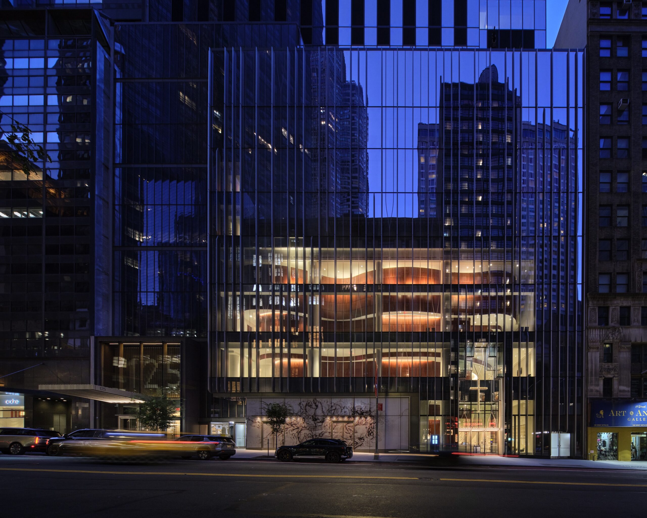 Modern glass building at dusk with illuminated interior and reflections of surrounding skyscrapers; reminiscent of the sleek design of the Lucas Museum of Narrative Art, with cars and light trails visible on the street in front.