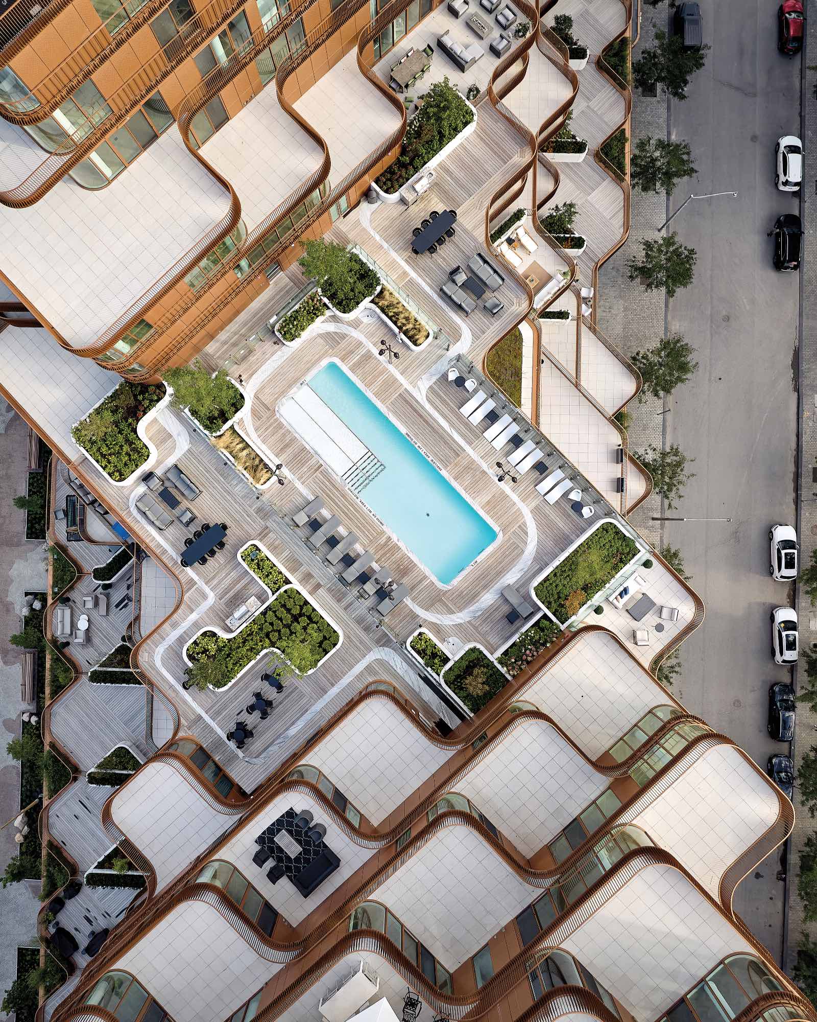 Aerial view of a modern building by 3XN with a central outdoor pool, surrounded by lounge chairs, greenery, and geometric architectural features; cars are parked on the adjacent street.