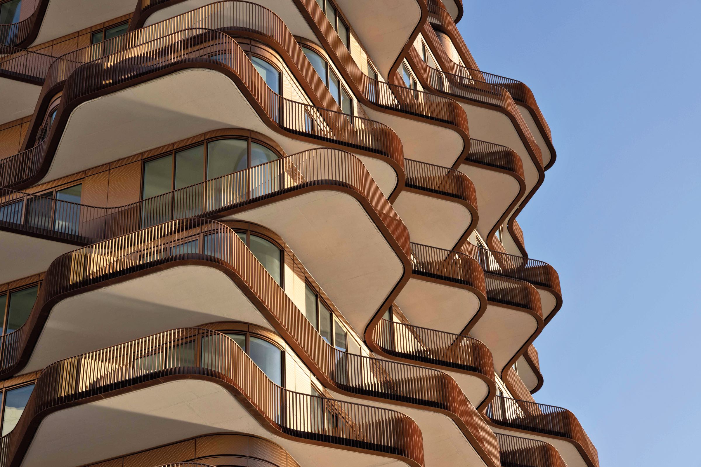 Curved balconies by 3XN with brown railings and glass windows on a modern building facade, viewed from below against a clear blue sky.