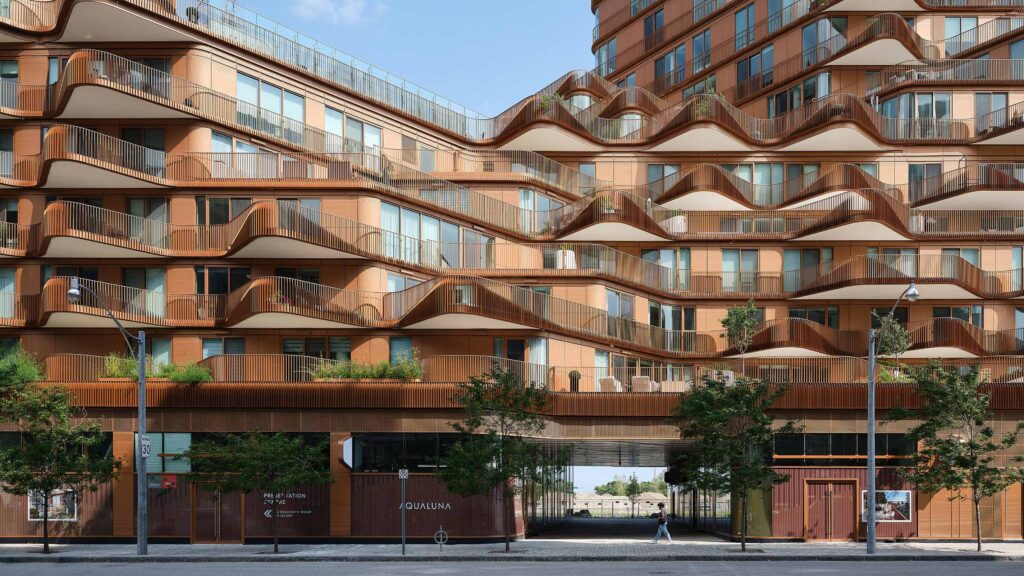 Modern apartment building by 3XN with wavy balconies, large windows, ground-floor shops, and trees lining the sidewalk.