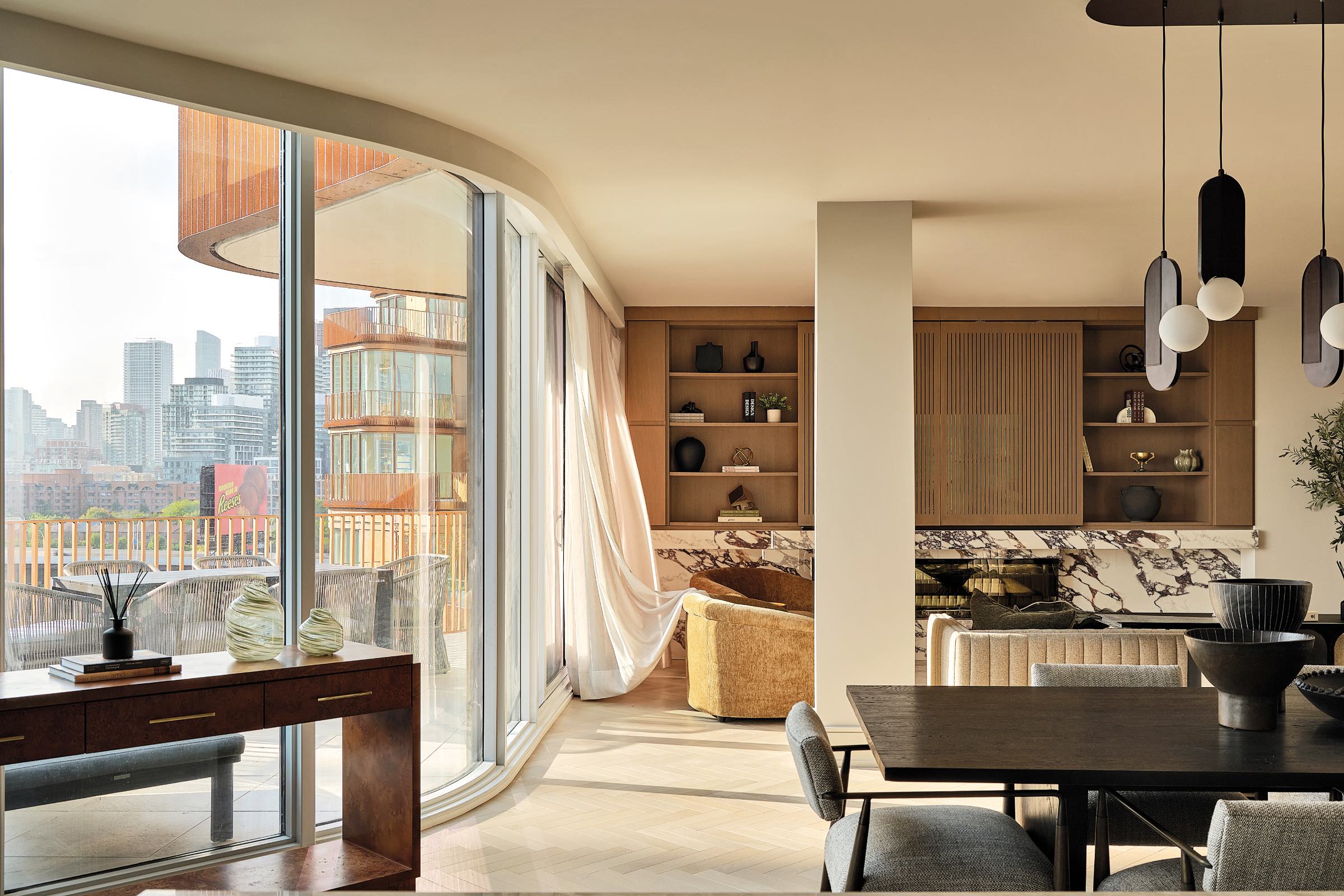Modern apartment interior with large windows, dining area, built-in shelves, marble accents, and city skyline views. Natural light fills the room, highlighting contemporary decor and furnishings.