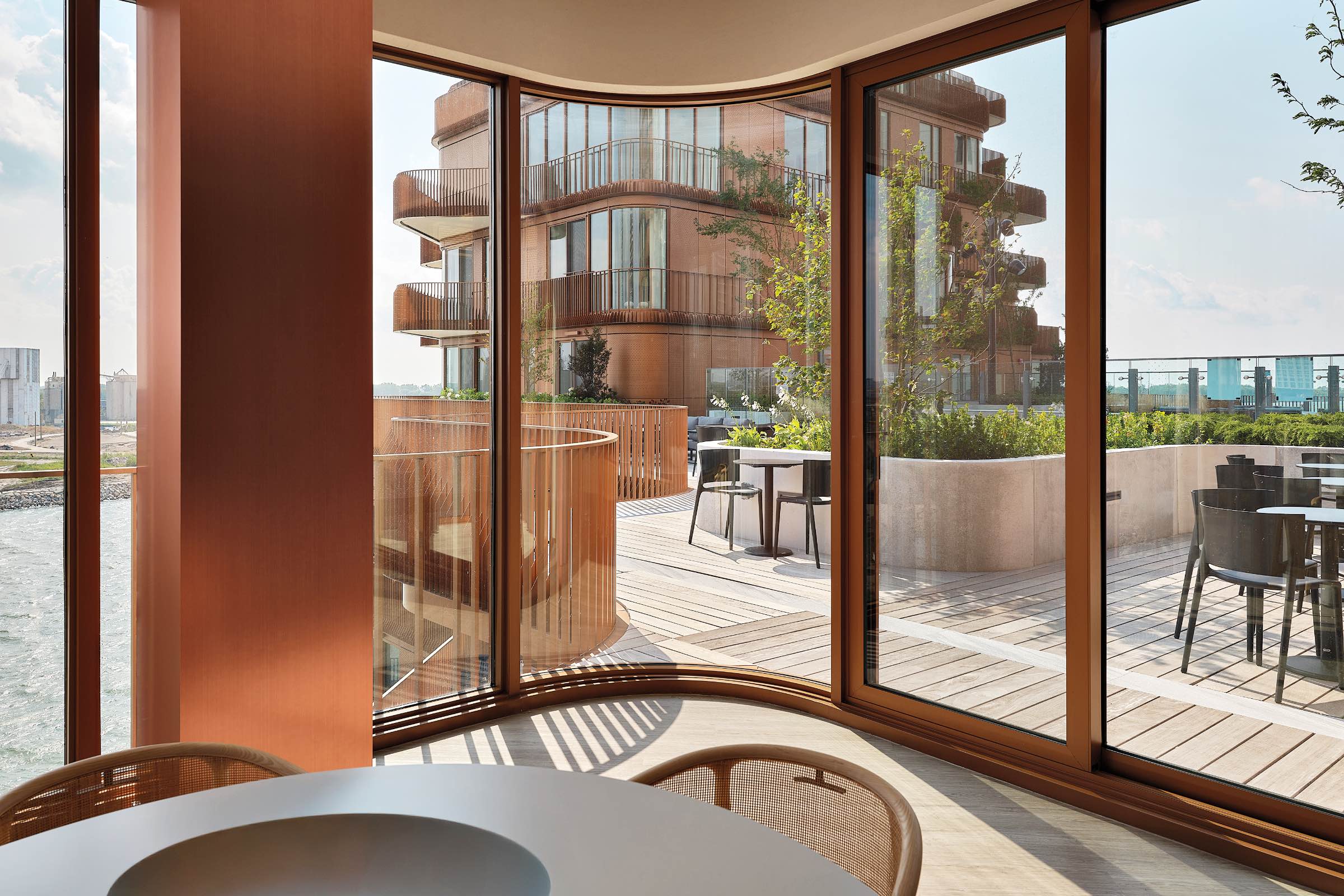Modern indoor dining area with curved glass walls overlooks an outdoor wooden deck, seating area, and a contemporary multi-story building. Cityscape visible in the background.