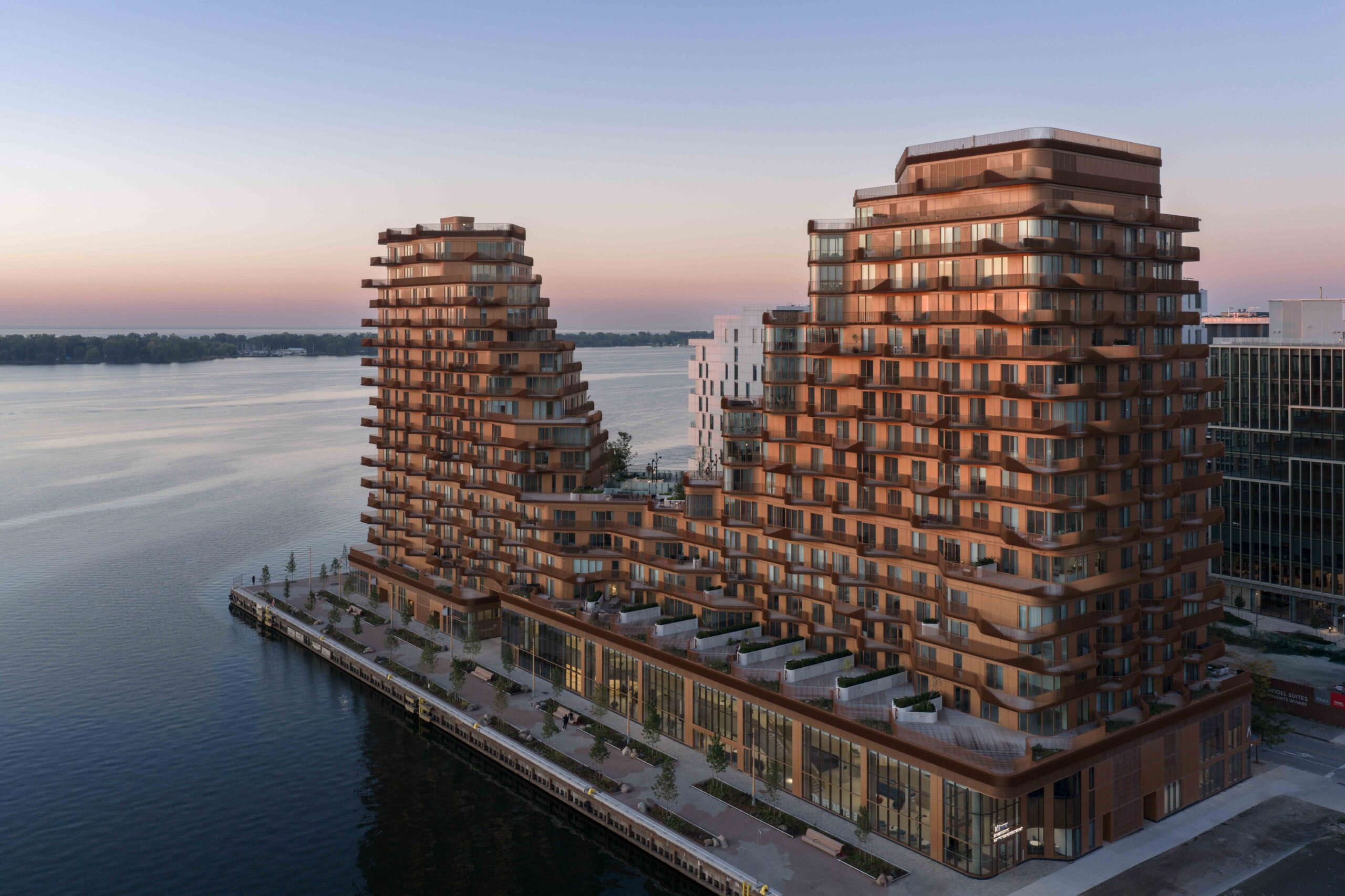 Two modern, multi-story buildings by 3XN with tiered balconies overlook a waterfront at sunset. The buildings feature warm-toned facades and are surrounded by walkways and water.
