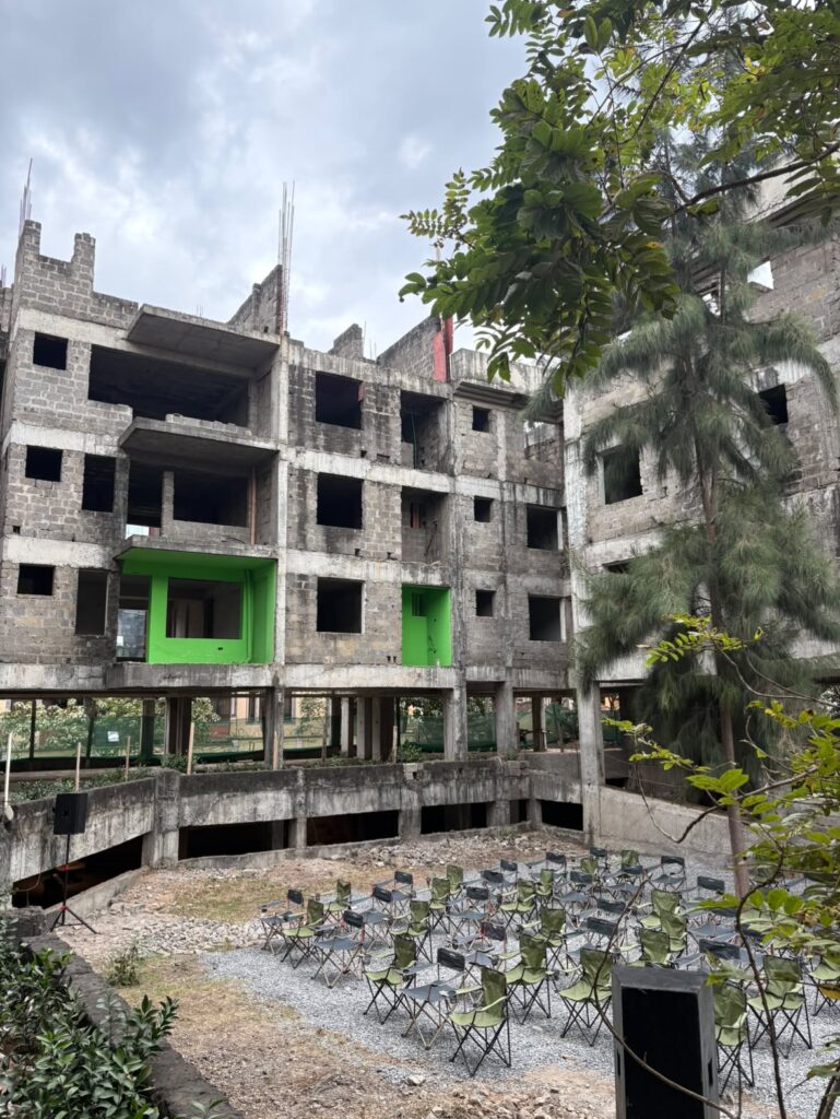 An unfinished concrete building with some green walls overlooks a courtyard where rows of folding chairs are arranged facing a small stage—an inviting scene reminiscent of gatherings planned for UIA 2026 Barcelona. Trees and a cloudy sky are visible, adding to the atmosphere.