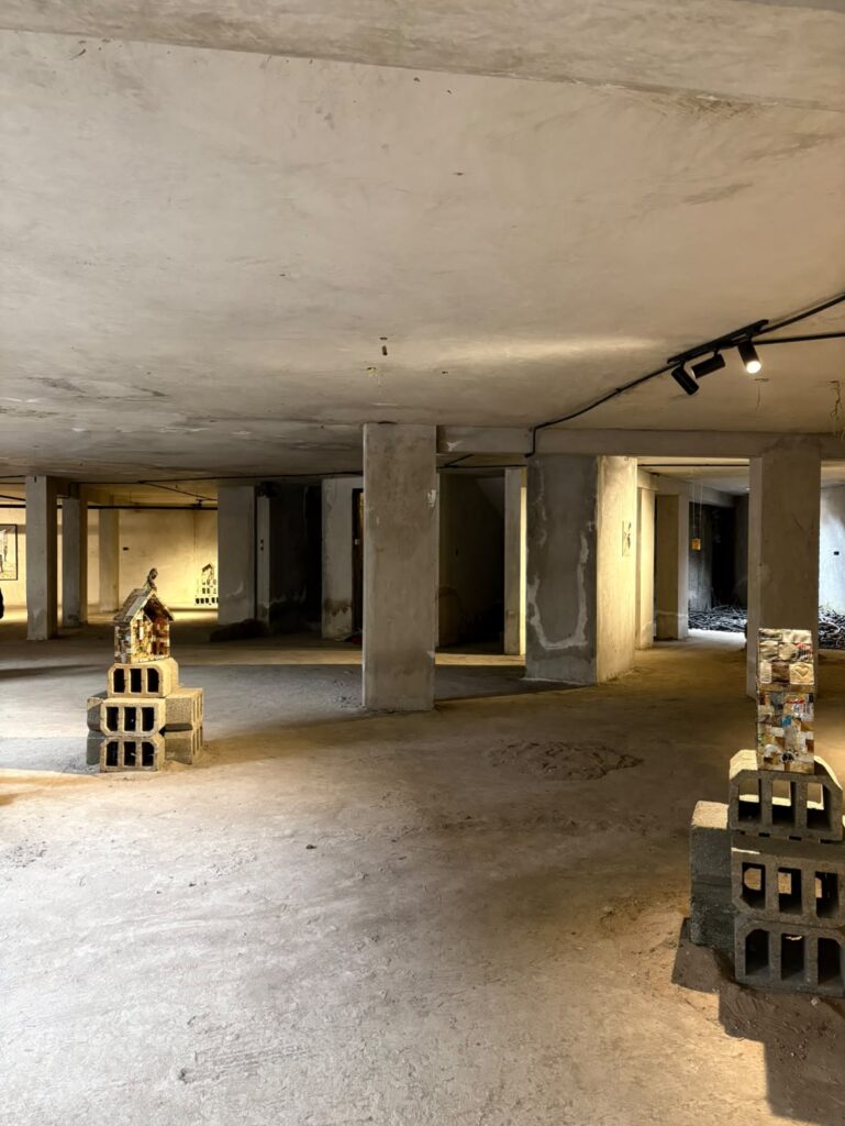 An unfinished concrete basement with several pillars, exposed ceilings, stacked cinder blocks, and small art displays arranged throughout the space creates an atmosphere reminiscent of the raw urban venues anticipated for UIA 2026 Barcelona.