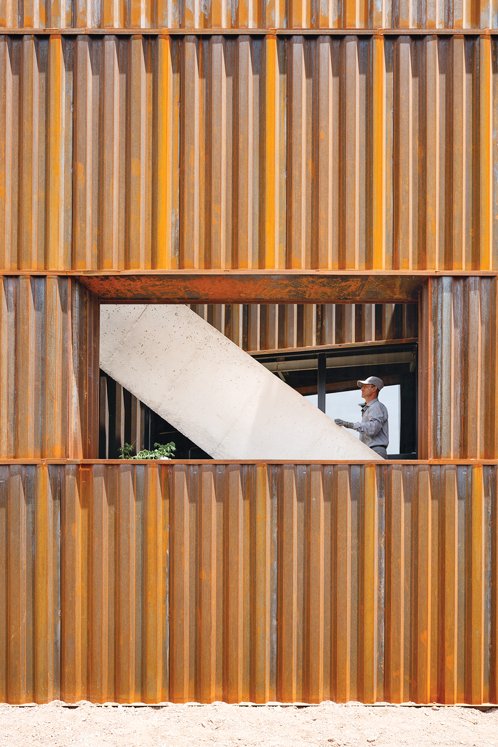 A person in a hard hat stands by a window inside a rust-colored corrugated metal building by Davood Boroojeni, next to a white diagonal structure.