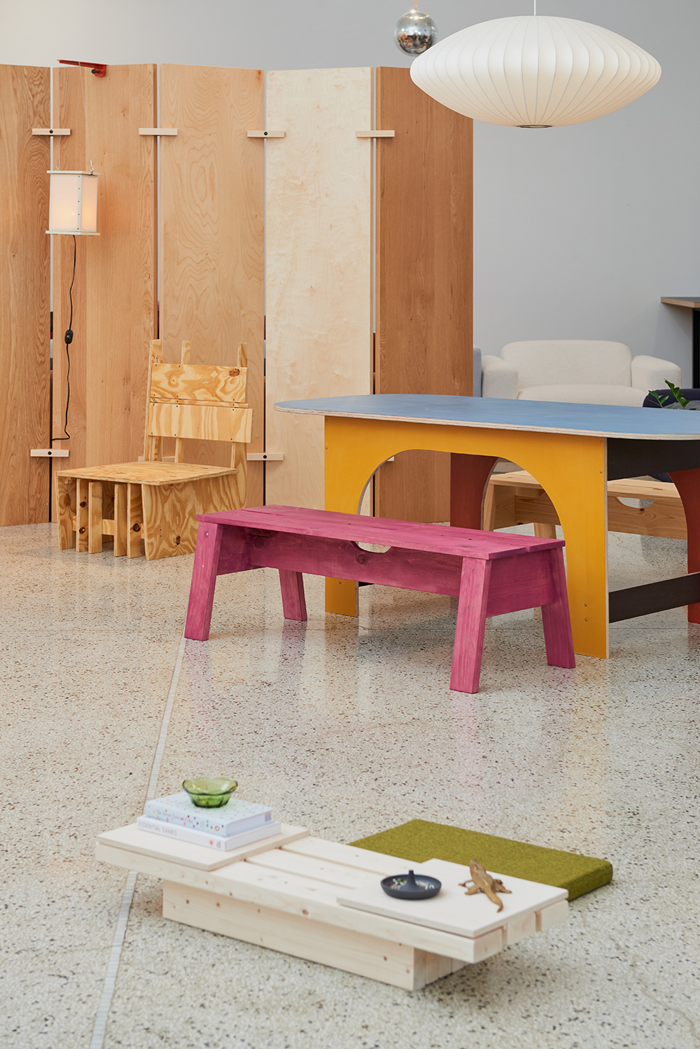 A room with a pink wooden bench, a blue and yellow table, light wood chairs, a low table with items, and a white pendant lamp.