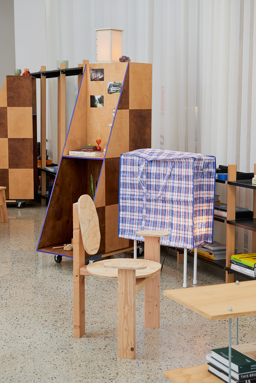 Wooden furniture and shelving units are arranged in a room with terrazzo flooring and sheer curtains; a blue plaid fabric covers a box-like structure.
