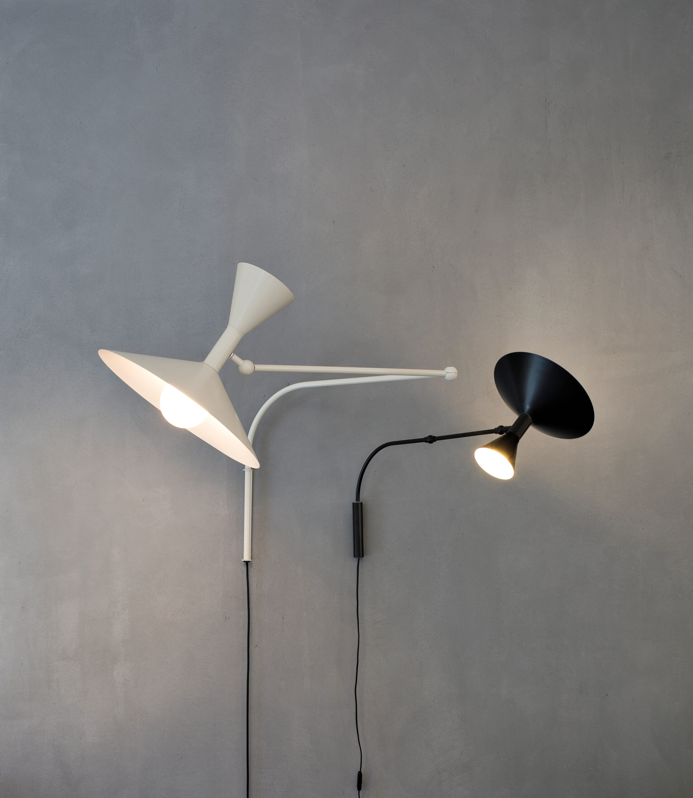 Products to Specify: Two wall-mounted lamps, one white and one black, are attached to a grey wall. Both lamps are on and have adjustable arms extending from the mounting points.