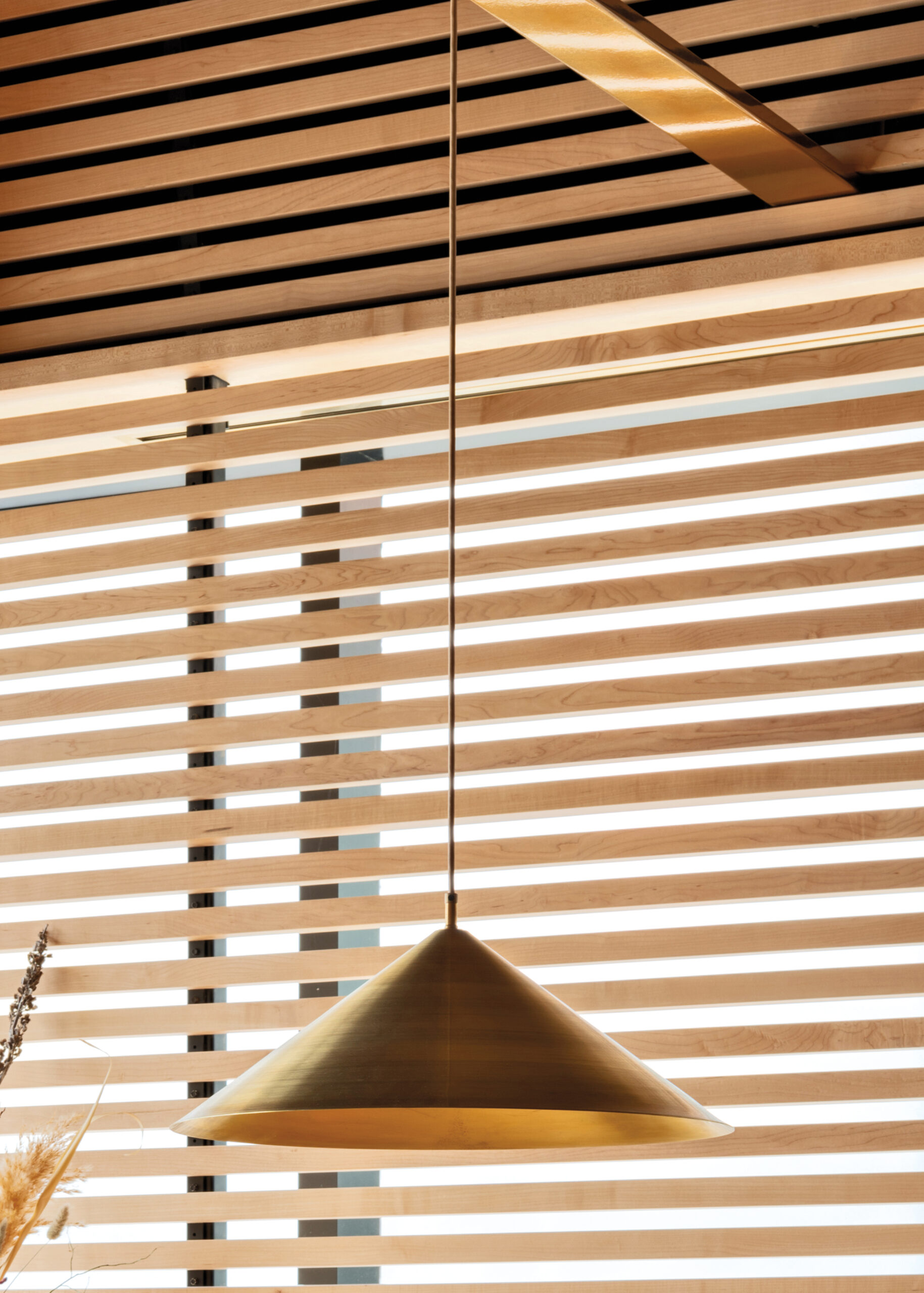 Products to Specify: A brass pendant light hangs from a slatted wood ceiling in front of horizontal wooden window blinds with sunlight streaming through.
