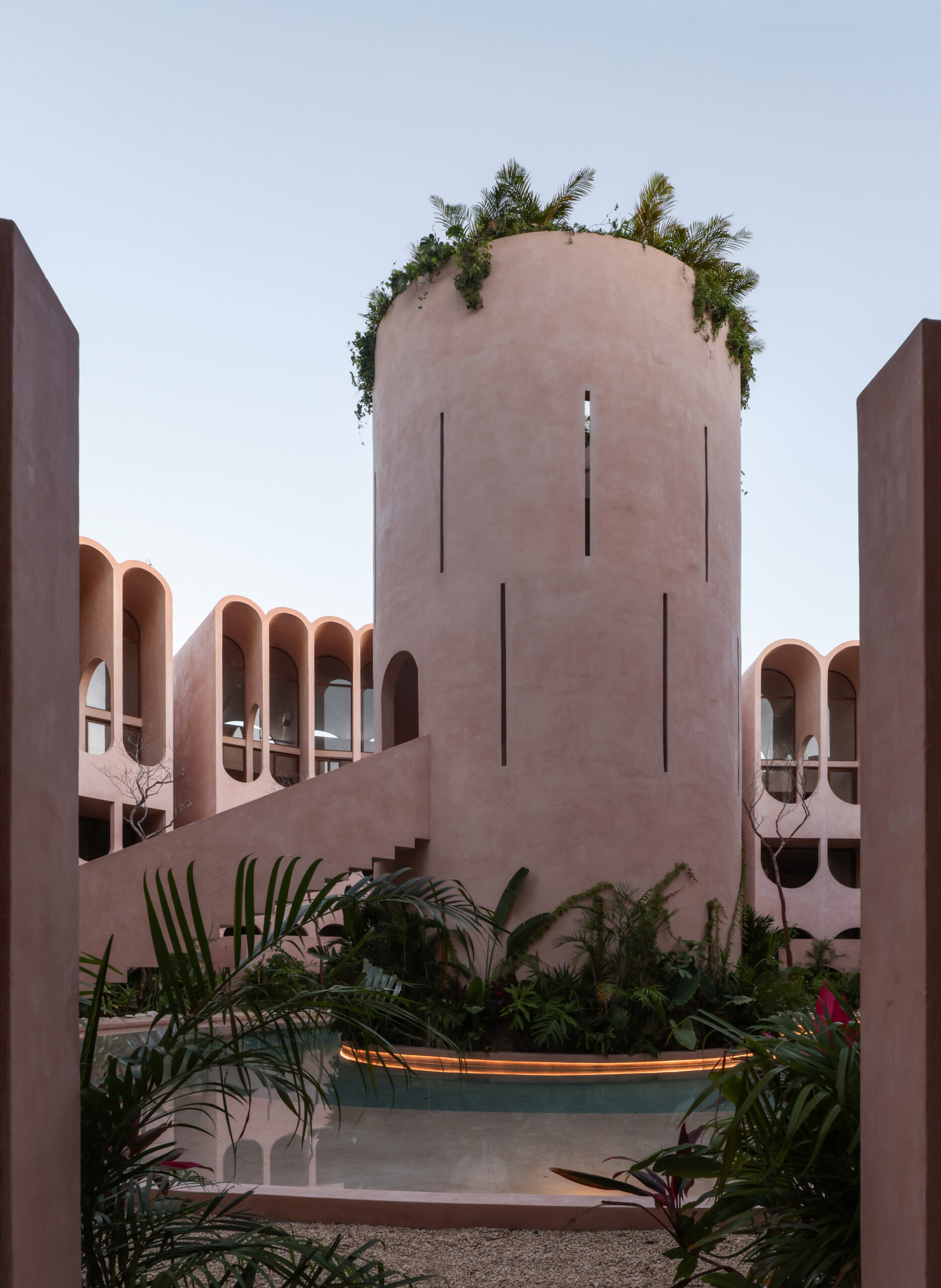 A light brown tower sits in the centre of a modern housing complex designed by V Taller in Tulum. Lush greenery and a reflective pool occupy the bottom of the frame, while a bright blue sky fills the top.