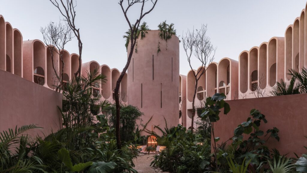 In Tulum, V Taller Inverts the Symbolism of the Tower of Babel A courtyard with pink stucco buildings, tall narrow windows, arched facades, sparse trees, and lush green plants in the foreground.