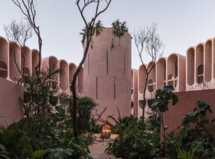 In Tulum, V Taller Inverts the Symbolism of the Tower of Babel A courtyard with pink stucco buildings, tall narrow windows, arched facades, sparse trees, and lush green plants in the foreground.