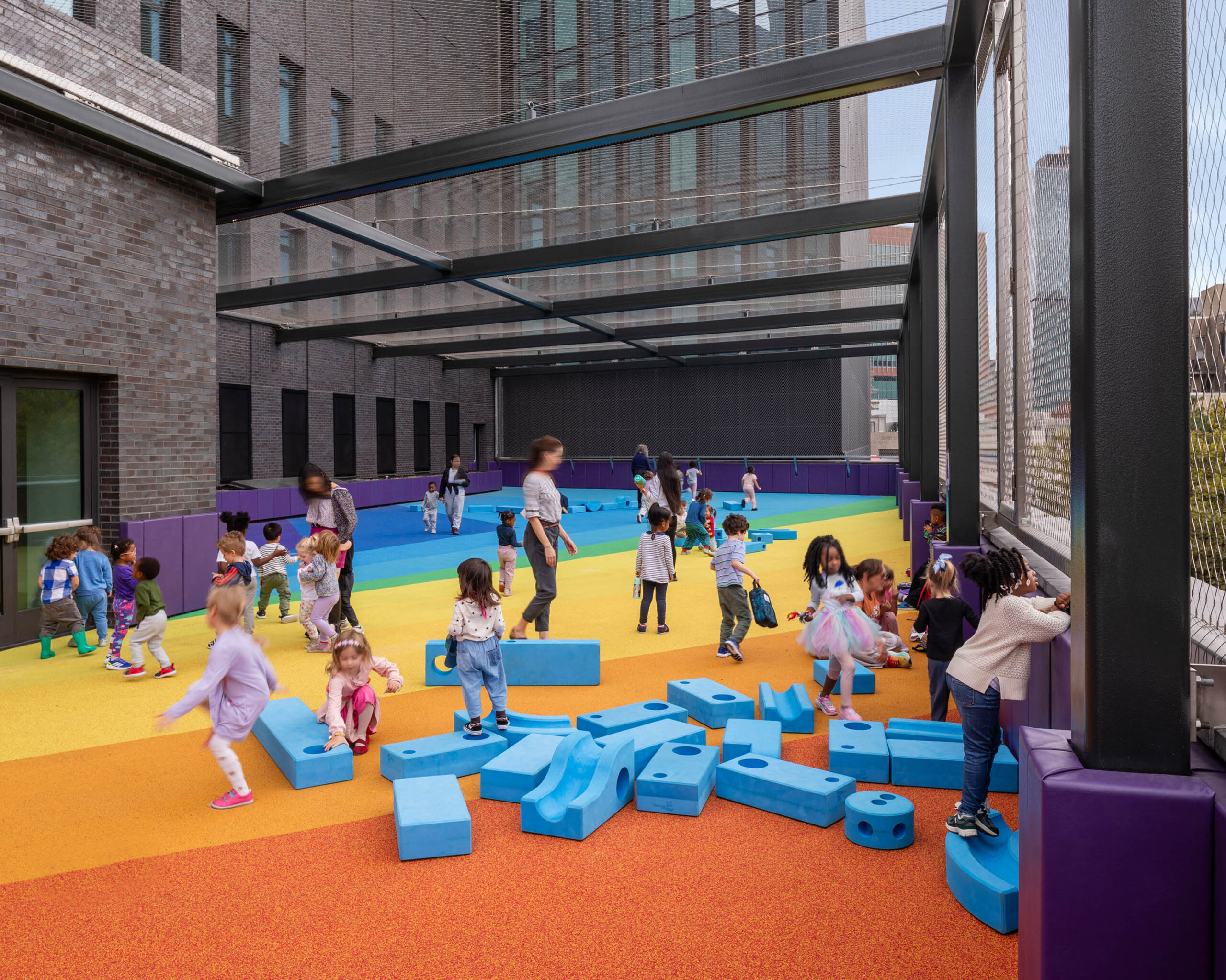 Children play on a colorful outdoor playground with foam blocks, surrounded by safety barriers and a netted enclosure, next to a brick building.