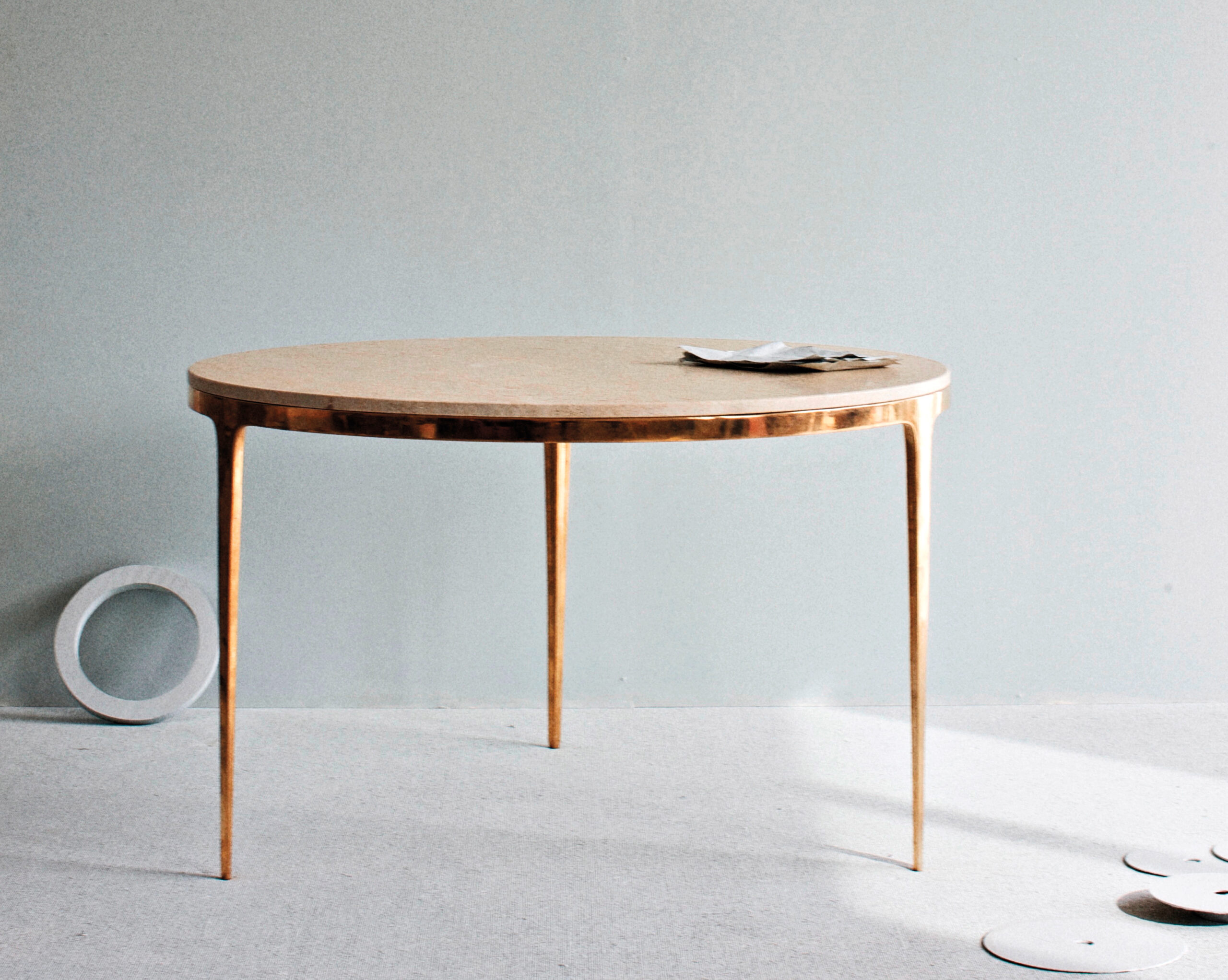 A round wooden table with thin gold legs stands on a light-colored floor. A few papers and objects are placed on top and beside the table.