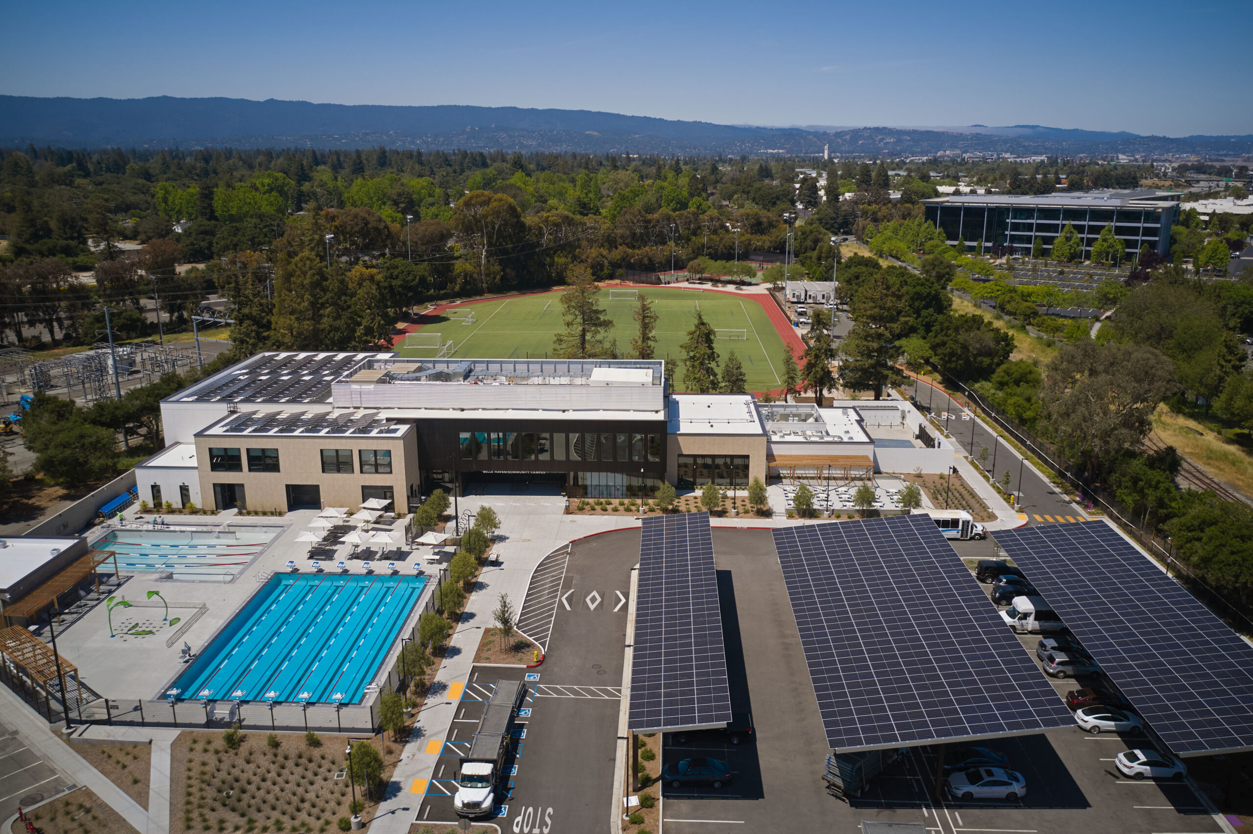 Aerial view of a modern campus with a swimming pool, solar panel-covered parking, athletic field, and surrounding trees under a clear sky.