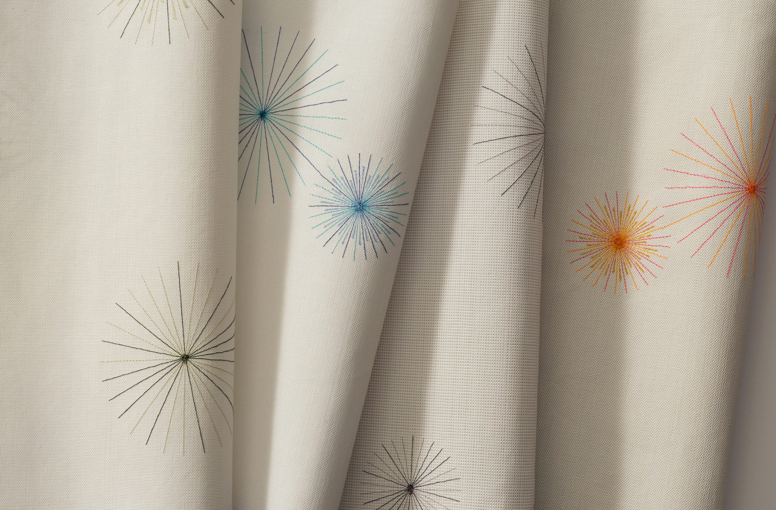 Close-up of cream-colored fabric with embroidered starburst patterns in various colors, including blue, orange, and green. The fabric is draped in soft folds.