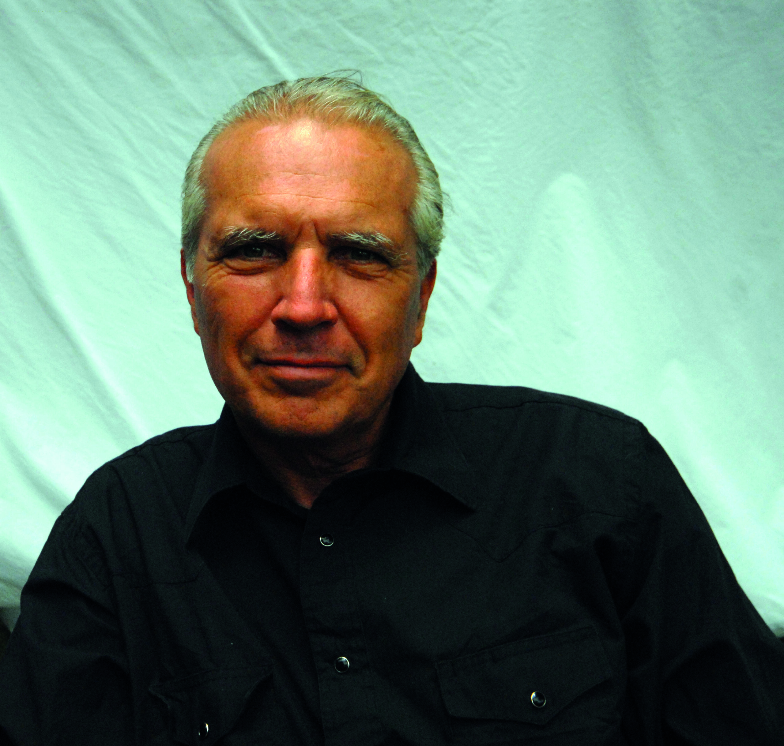 An older man with gray hair, wearing a black button-up shirt, sits in front of a light-colored fabric backdrop, looking at the camera with a neutral expression.