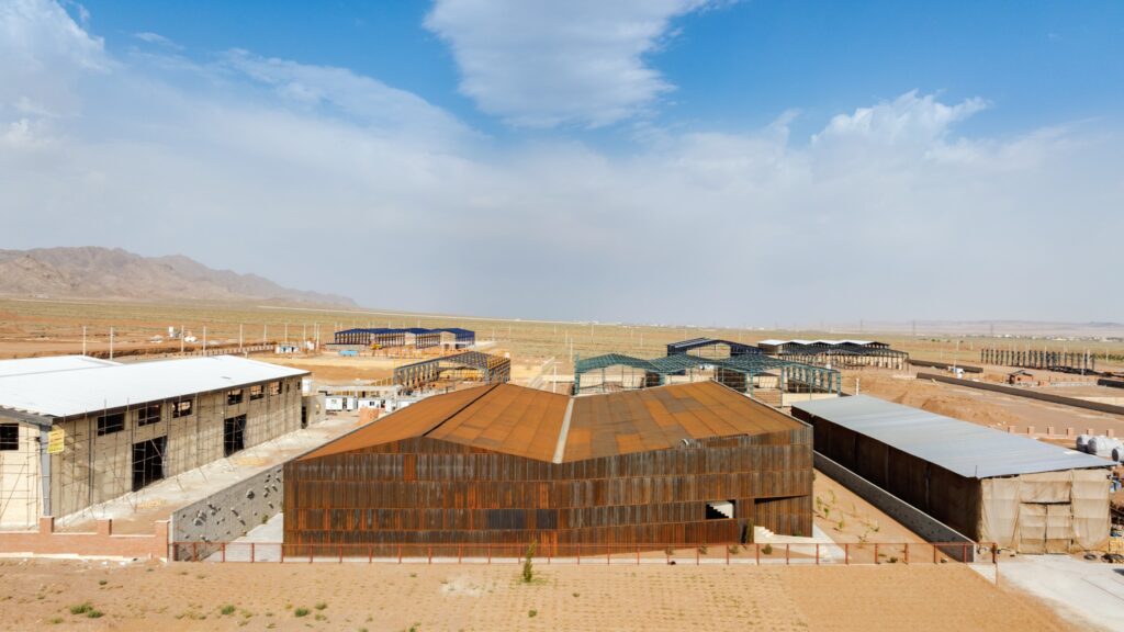 Several industrial buildings and greenhouses sit on a flat, arid landscape under a partly cloudy sky with distant mountains in the background.