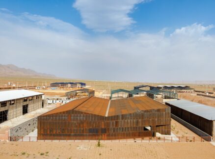 Several industrial buildings and greenhouses sit on a flat, arid landscape under a partly cloudy sky with distant mountains in the background.