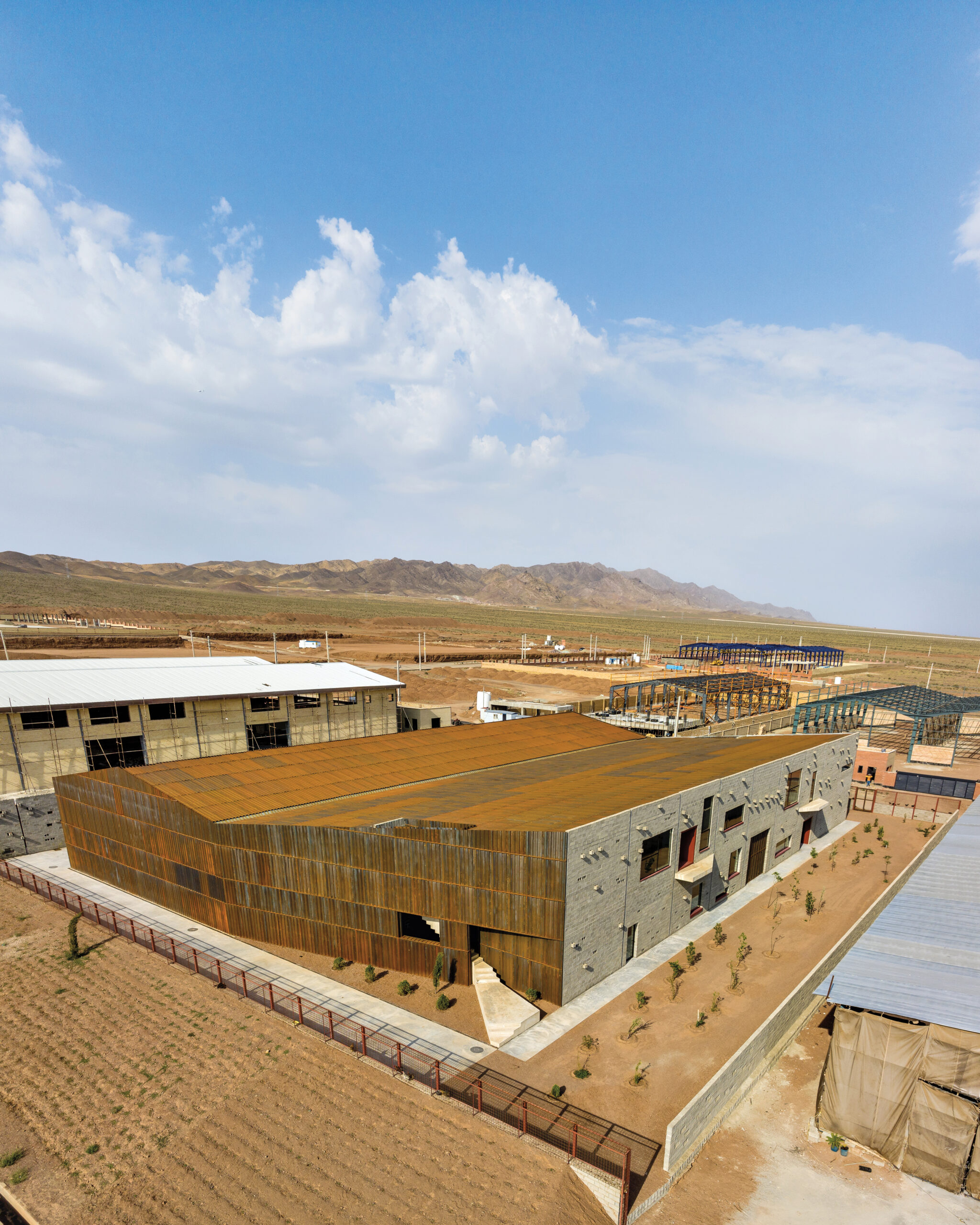 In a flat, arid landscape under a partly cloudy sky with distant mountains in the background, Davood Boroojeni's new factory building features rust-colored wall panels in an angular layout. Also visible are building frames and distant fields.