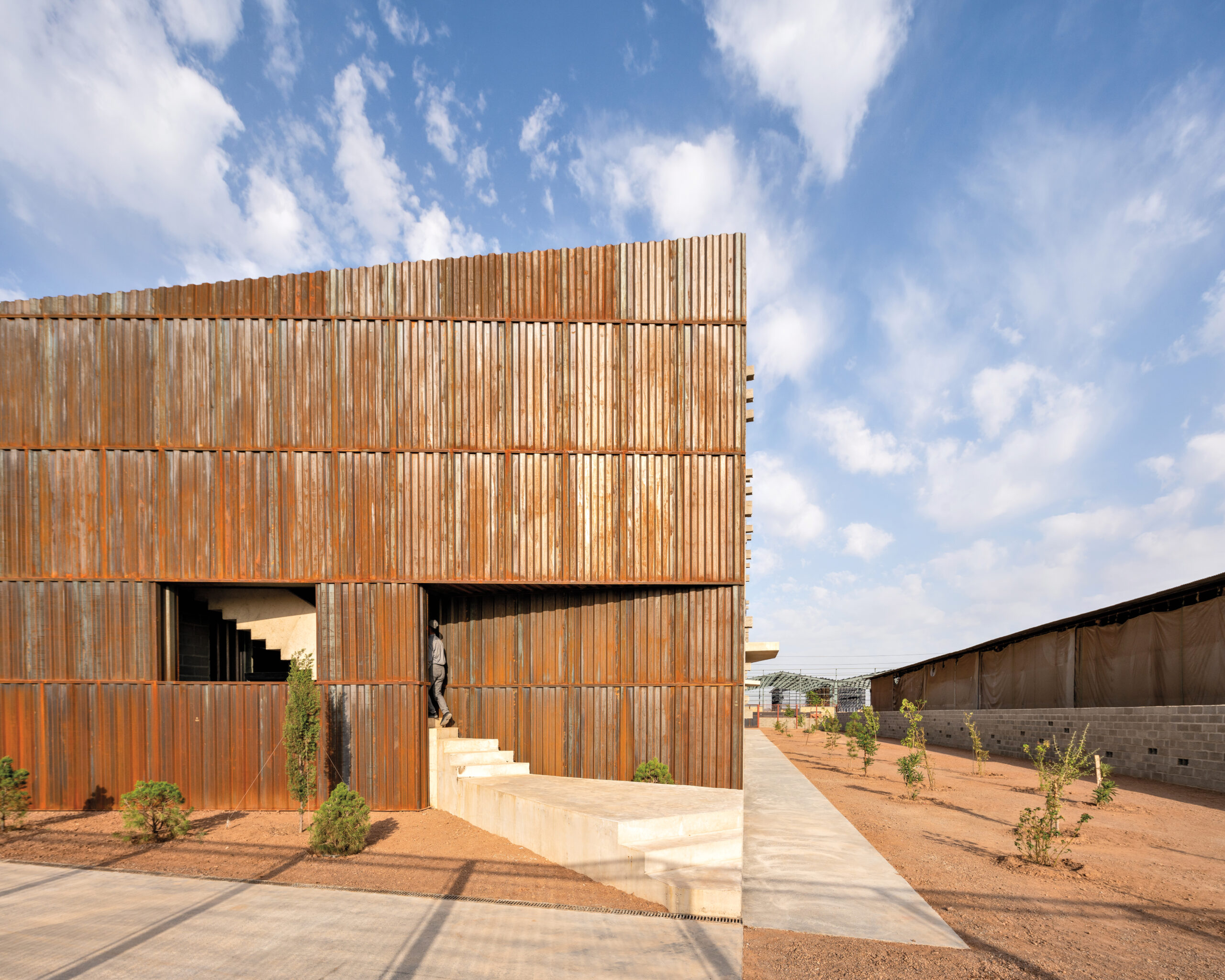 In a flat, arid landscape under a partly cloudy sky with distant mountains in the background, Davood Boroojeni's new factory building features rust-colored wall panels in an angular layout.