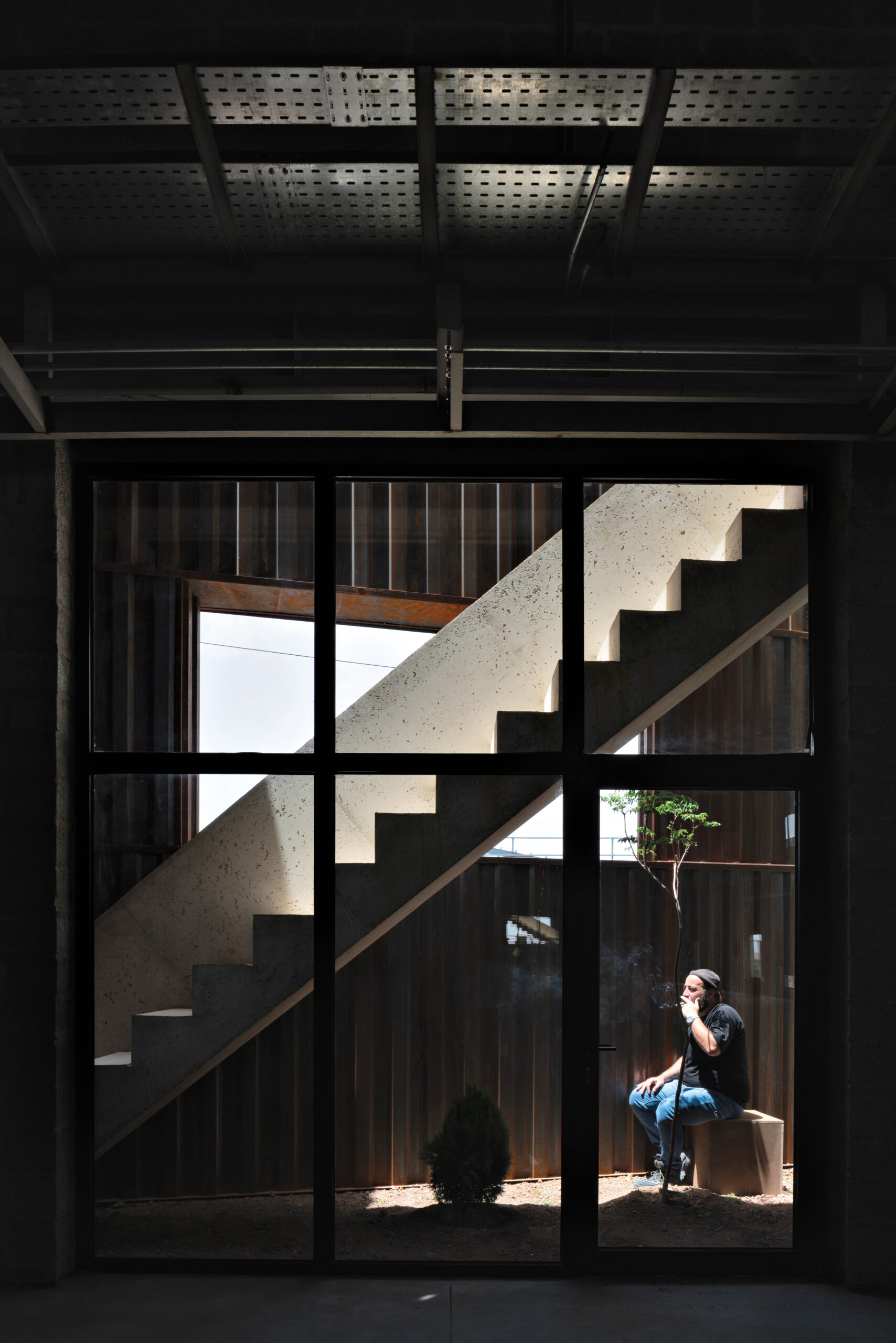 Inside of a darkly-lit factory with. a man seated in the bottom right, smoking a cigarette underneath a concrete staircase.