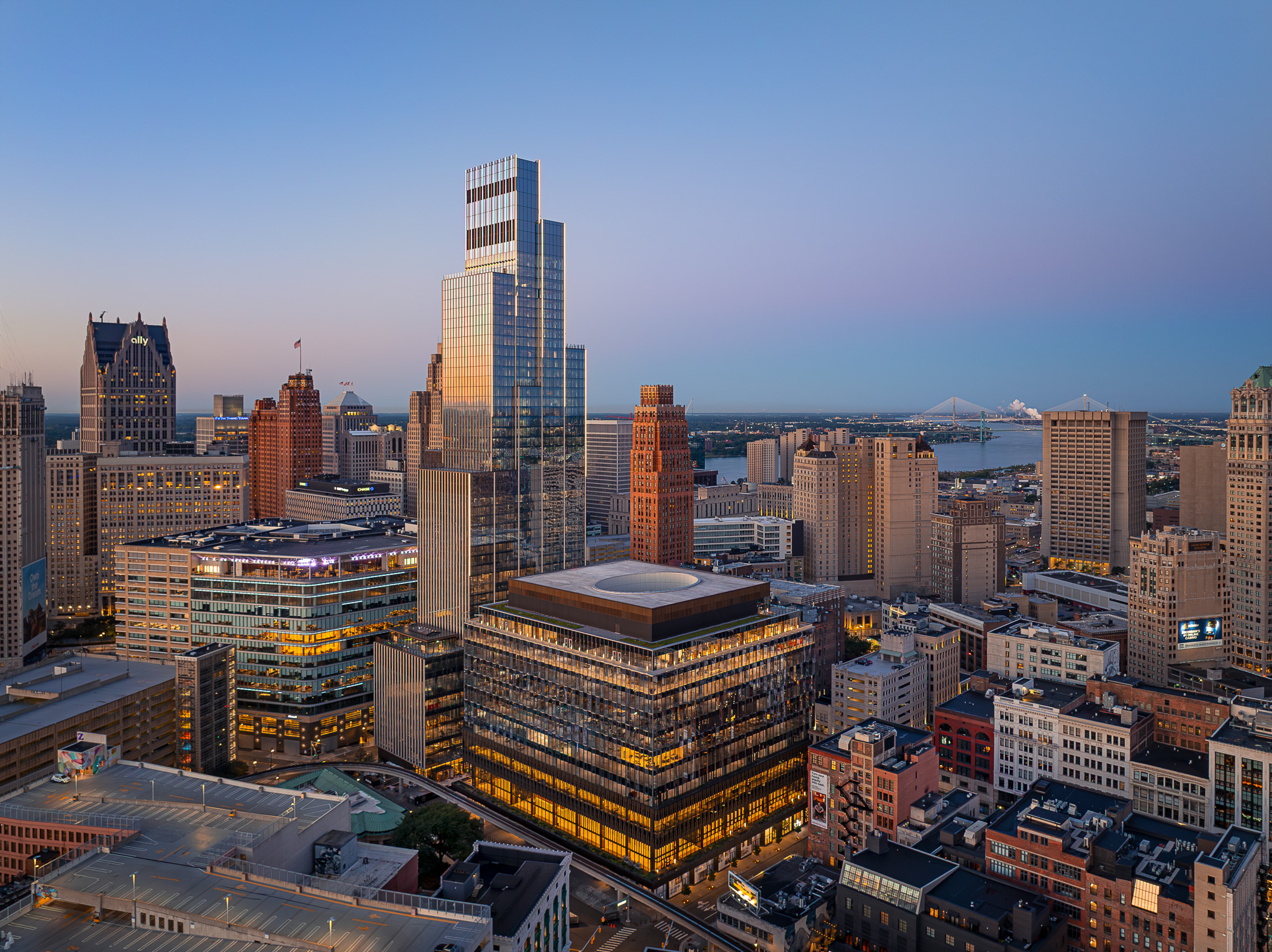 A cityscape at dusk shows modern and historic buildings in downtown Detroit, Michigan, with the sky transitioning from blue to pink near the horizon.