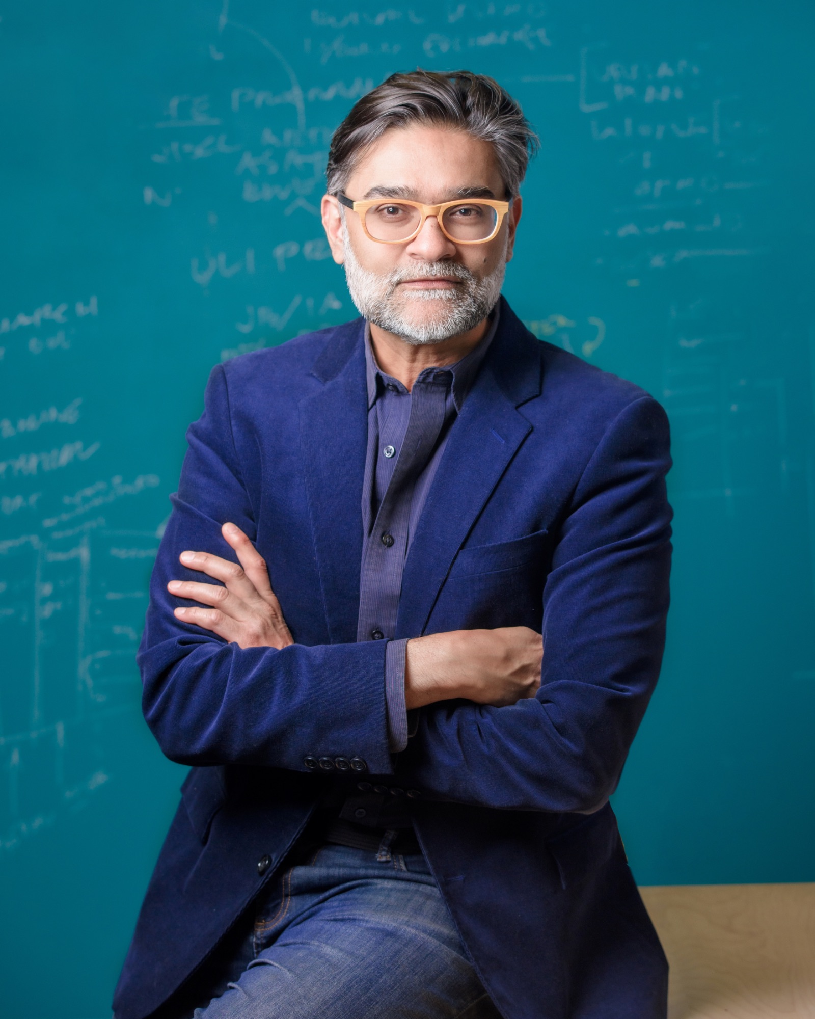 Man with gray hair and beard wearing glasses, a blue blazer, and jeans sits with arms crossed in front of a teal chalkboard with handwritten notes.