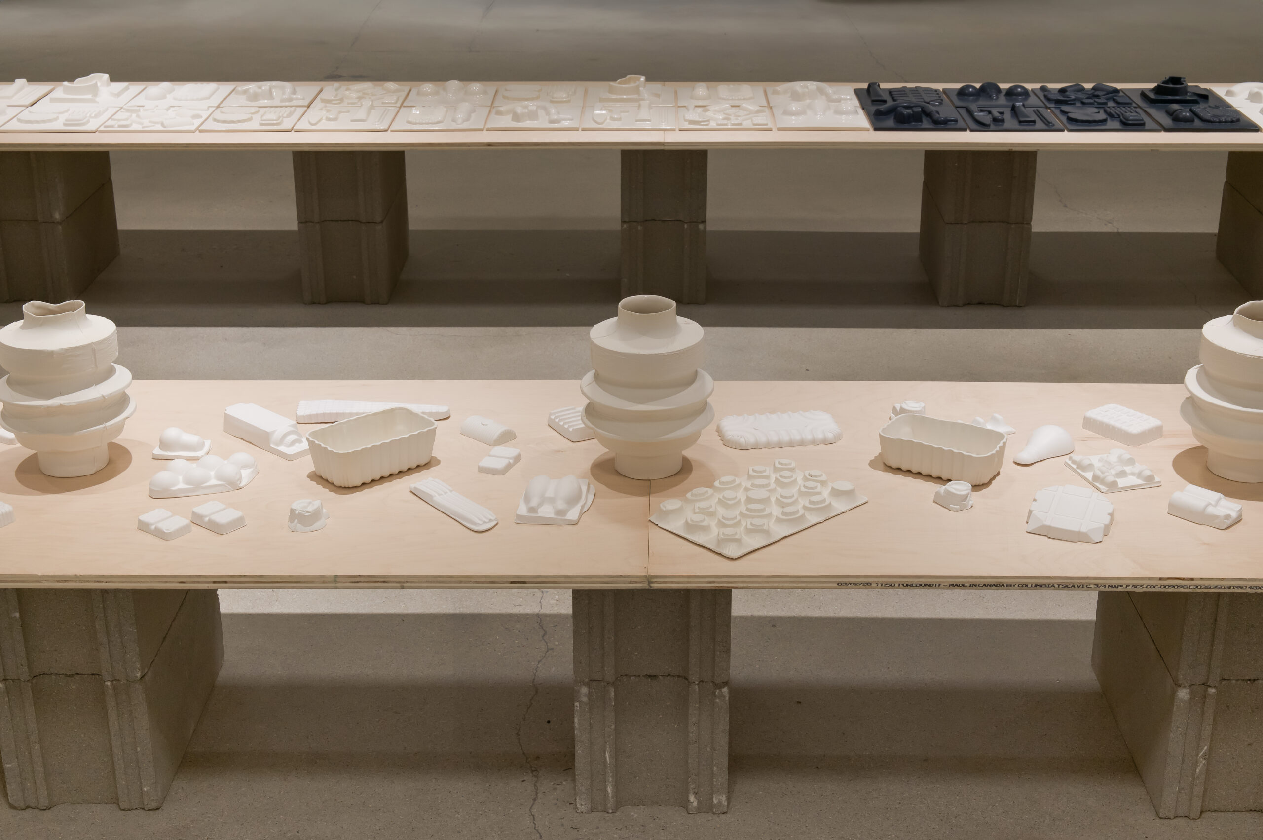 Tables display various white and black sculptural objects, including vases, trays, and geometric forms, arranged in orderly rows on wooden surfaces supported by concrete blocks.