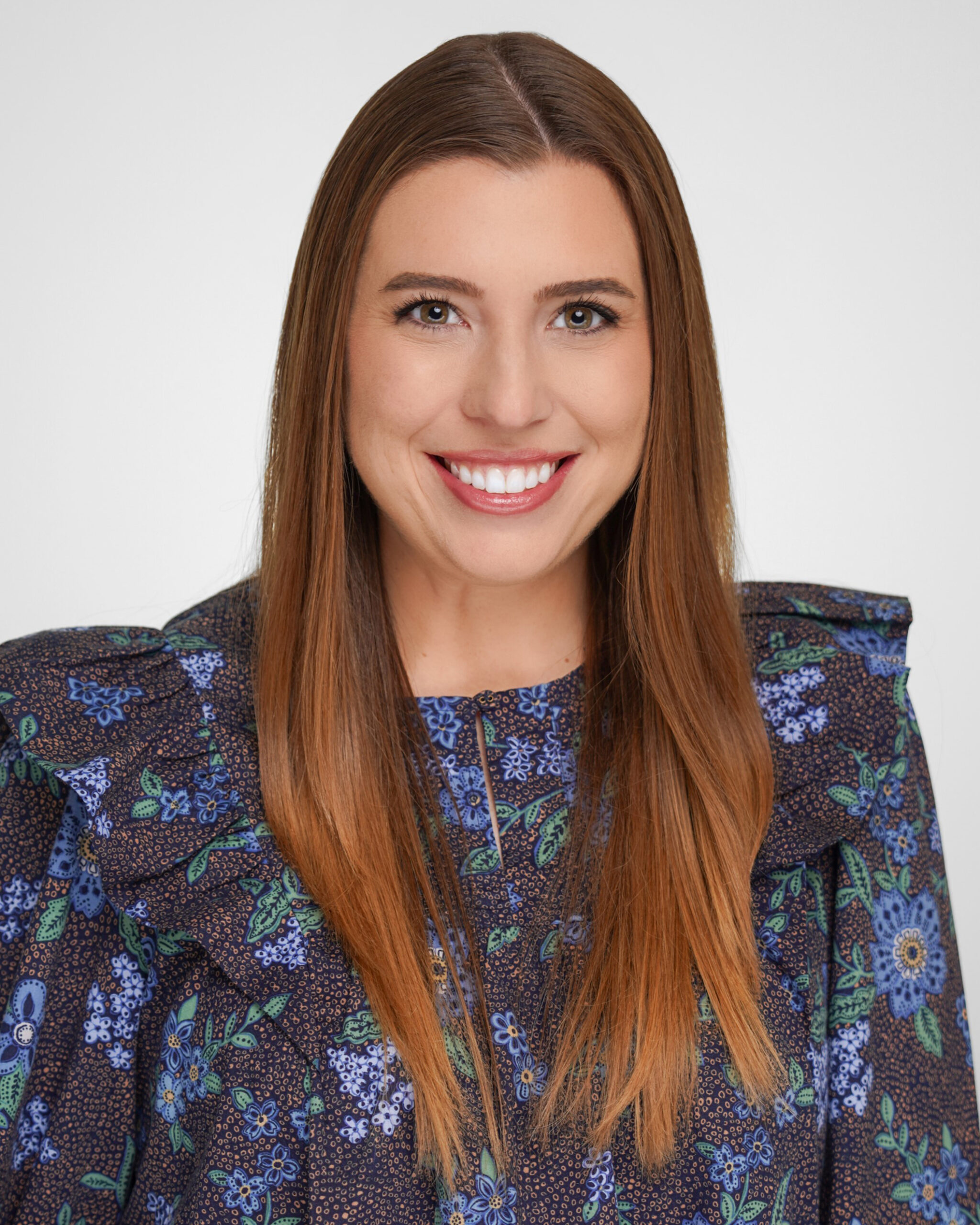 Woman with long straight brown hair, smiling, wearing a dark floral blouse with blue and purple flowers, posing in front of a plain light background.