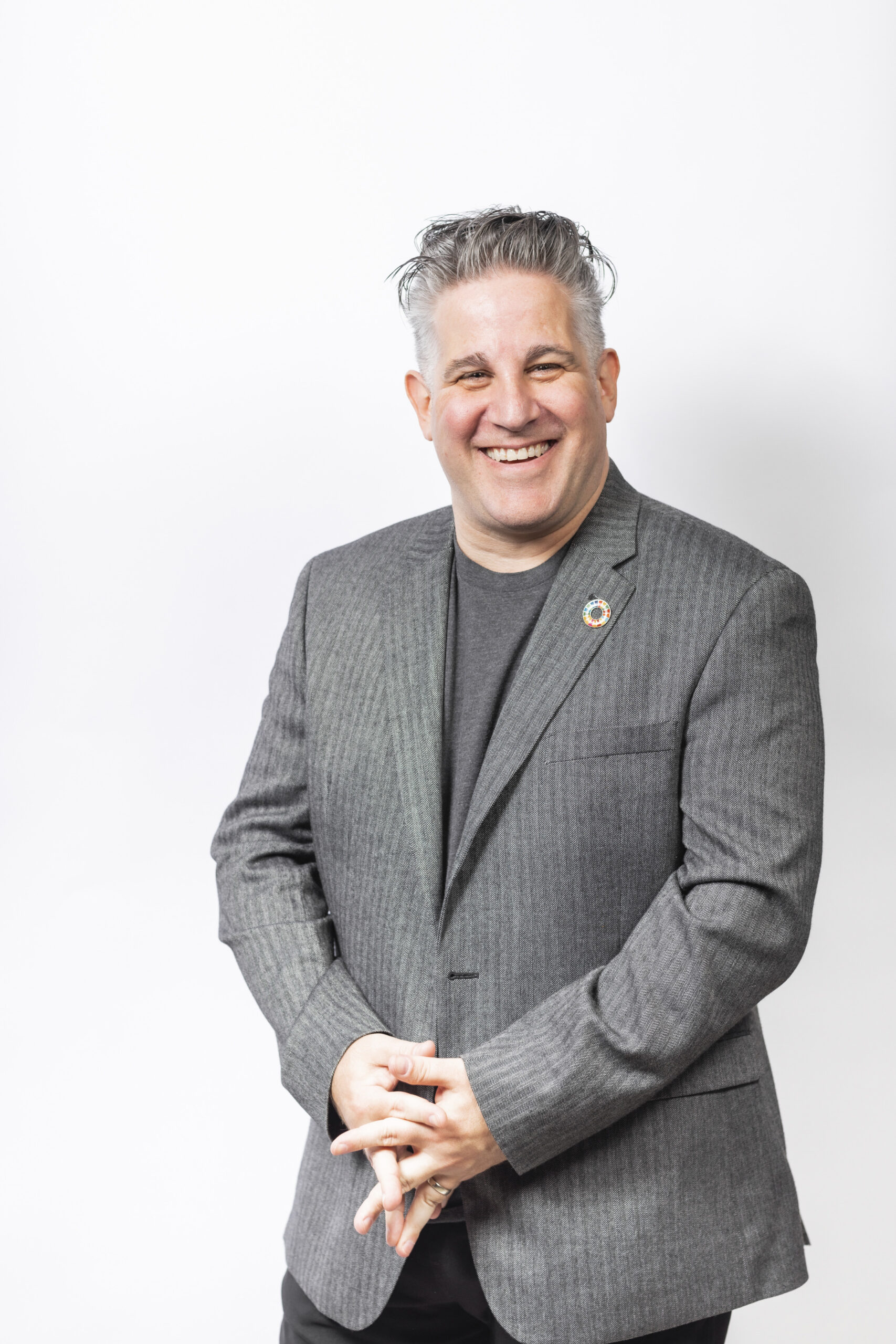 Smiling man with gray hair wearing a gray blazer and dark shirt stands against a plain white background, hands clasped in front.