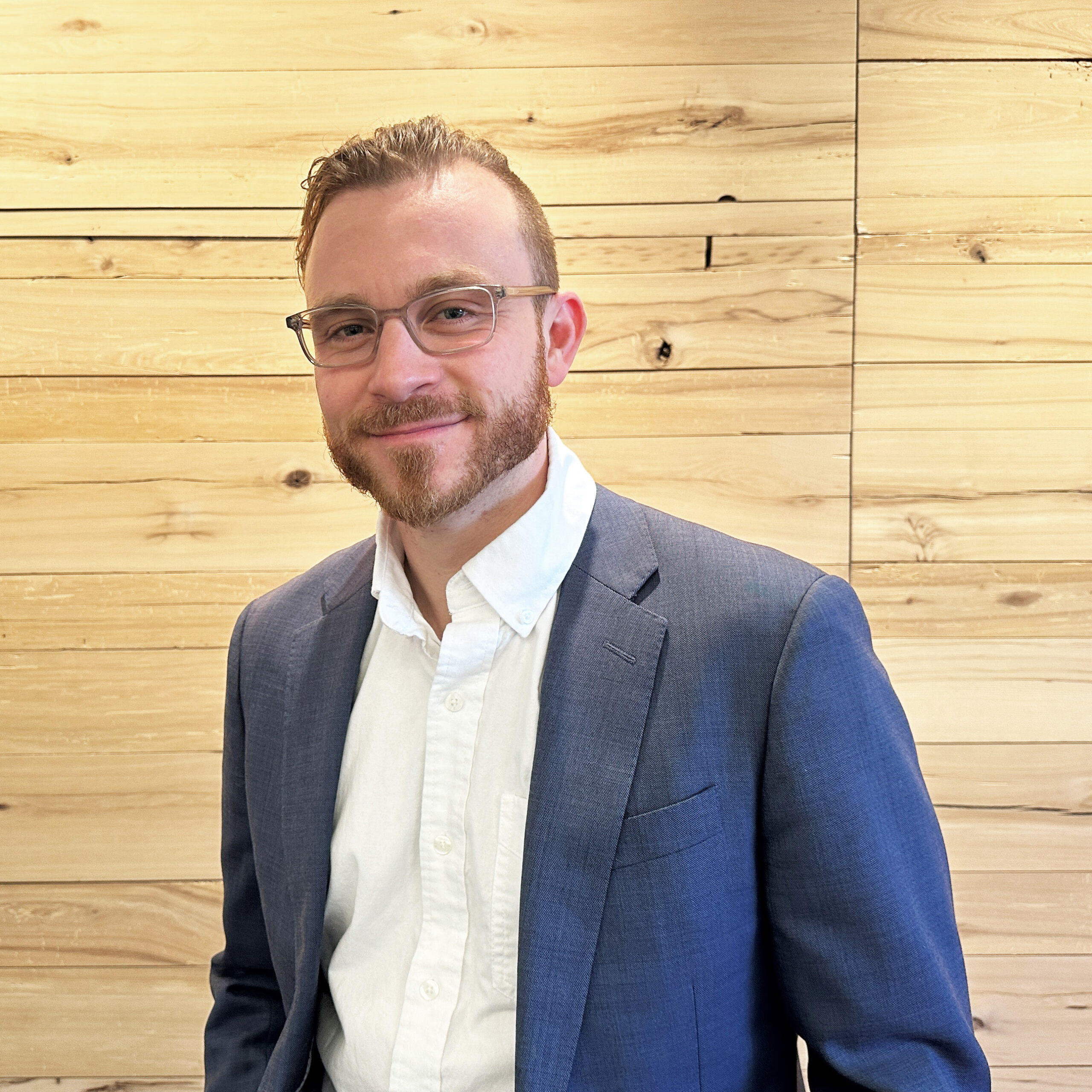 Man with glasses and a beard wearing a blue suit jacket and white shirt, standing in front of a wooden wall.