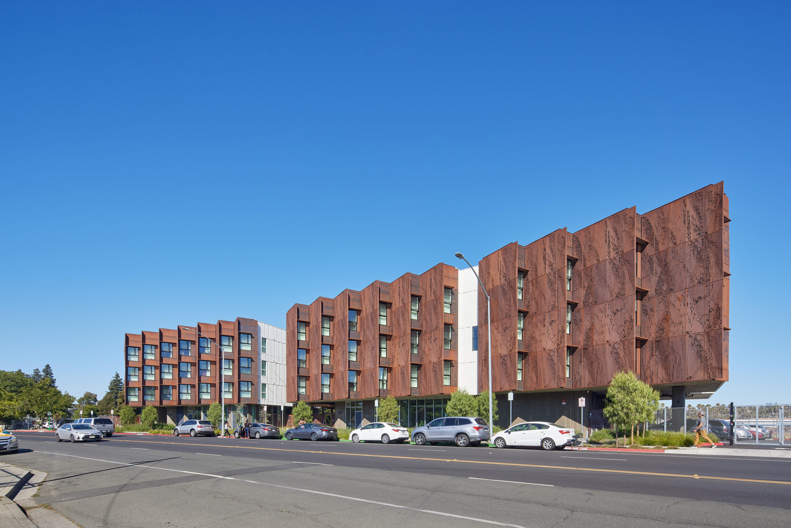 A modern four-story apartment building with rust-colored metal panels and parked cars along a sunny street.