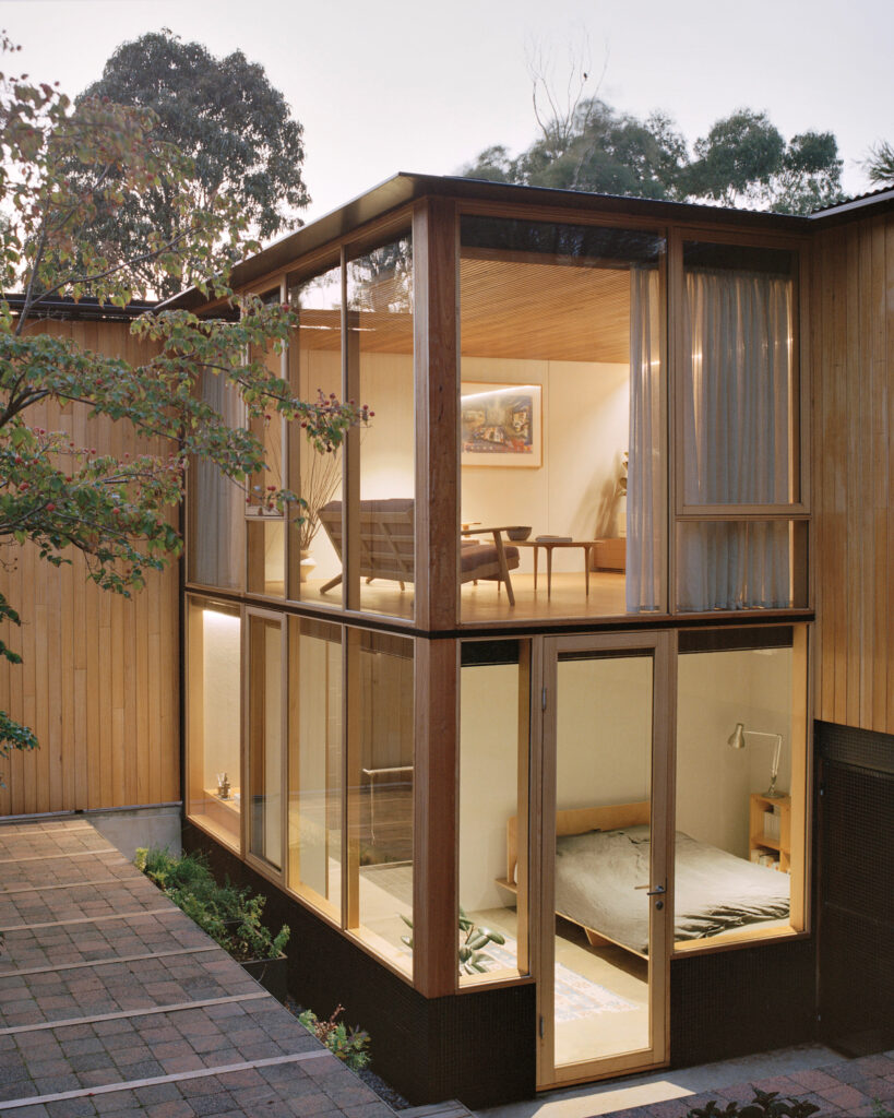 Products to Specify: Modern two-story house with large glass windows; upper level shows a furnished living area, lower level has a bedroom with a bed and nightstand; exterior features wood paneling and patio tiles.