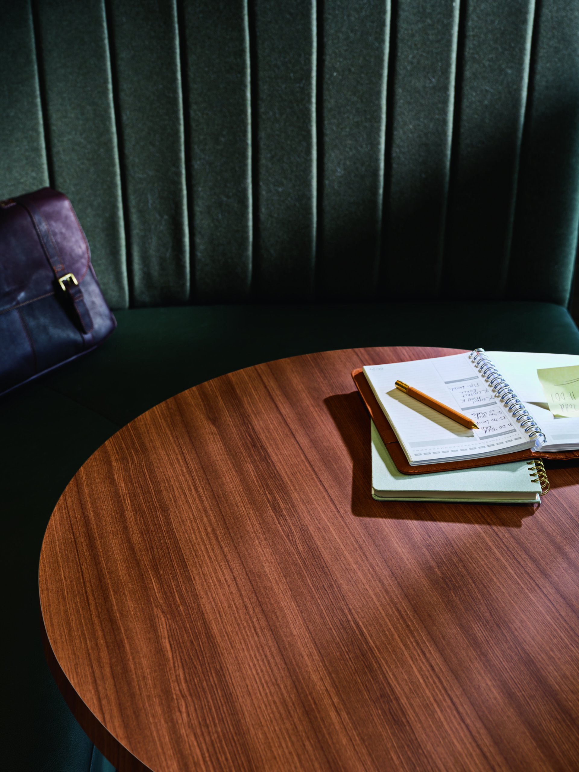 A wooden table with an open notebook, pencil, sticky notes, and a closed bag on a bench seat with vertical upholstery.