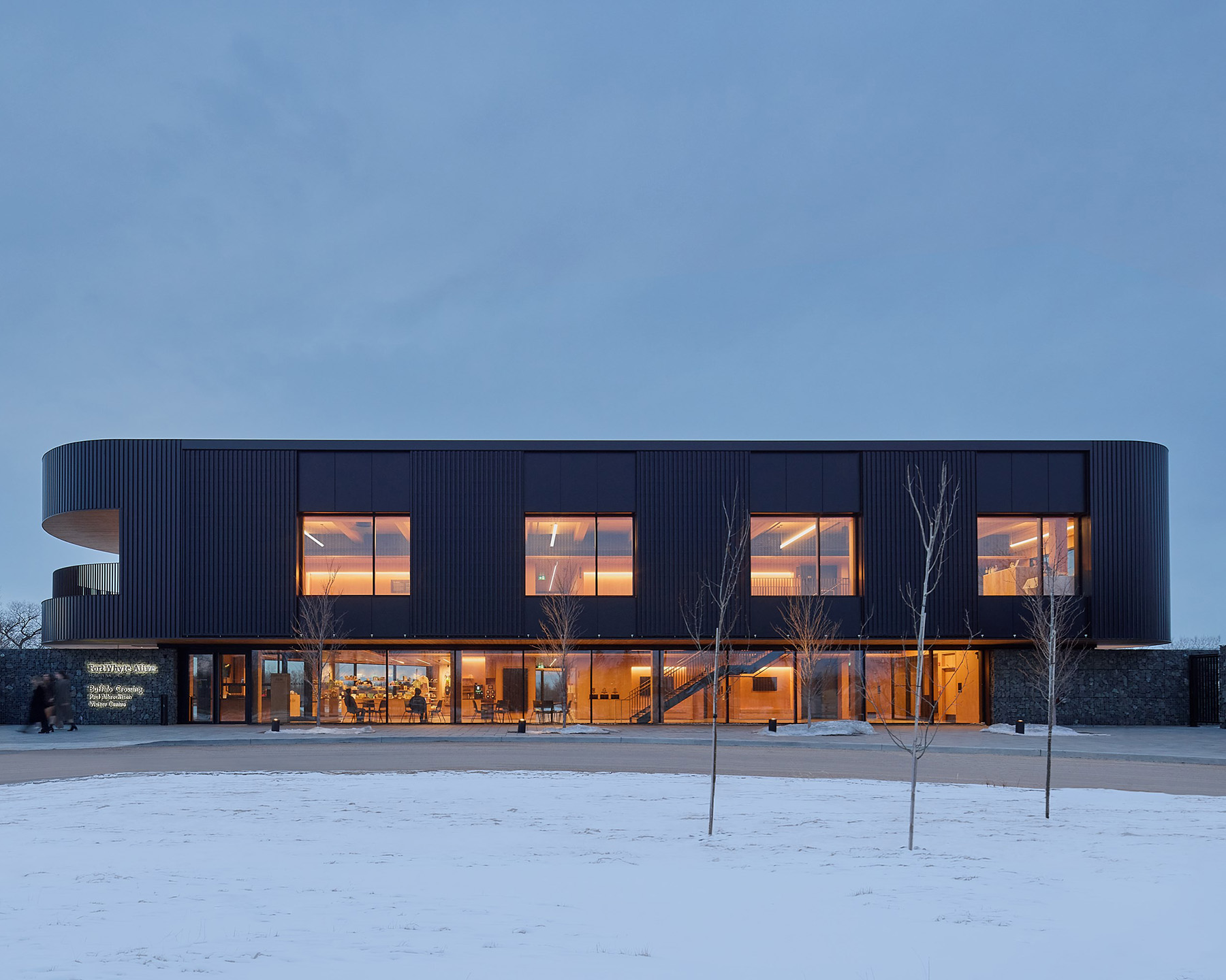Modern two-story building with large illuminated windows, dark exterior panels, and a snowy landscape in front.