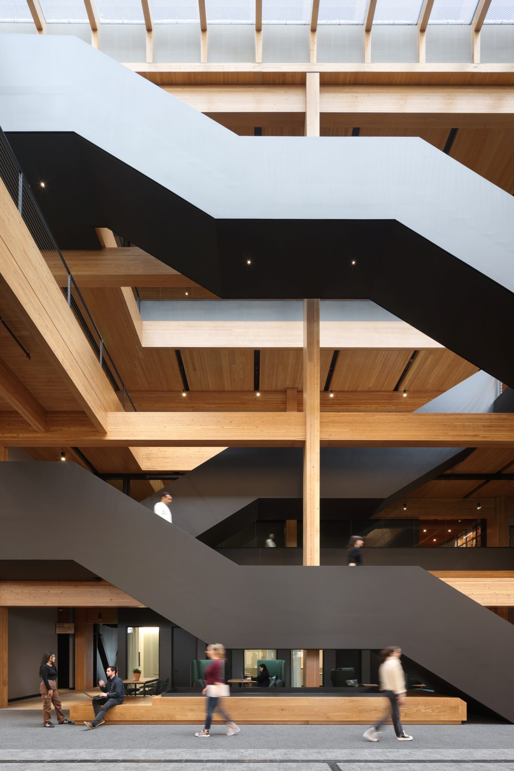Modern multi-story interior with wood beams, black angular staircases, a glass ceiling, and people walking and sitting throughout different levels—a space reminiscent of Michael Green’s innovative approach to architectural design.