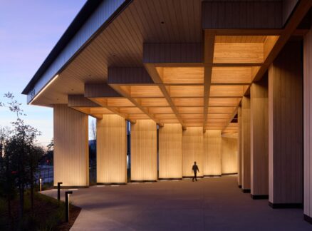 A person walks under a modern building designed by Michael Green, featuring tall, illuminated wooden columns and a geometric ceiling near dusk.