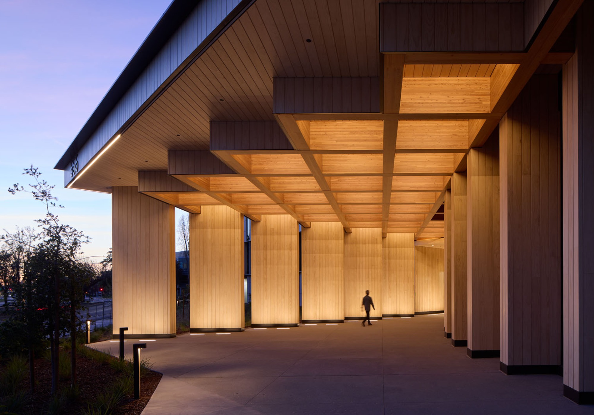 A person walks under a modern building designed by Michael Green, featuring tall, illuminated wooden columns and a geometric ceiling near dusk.