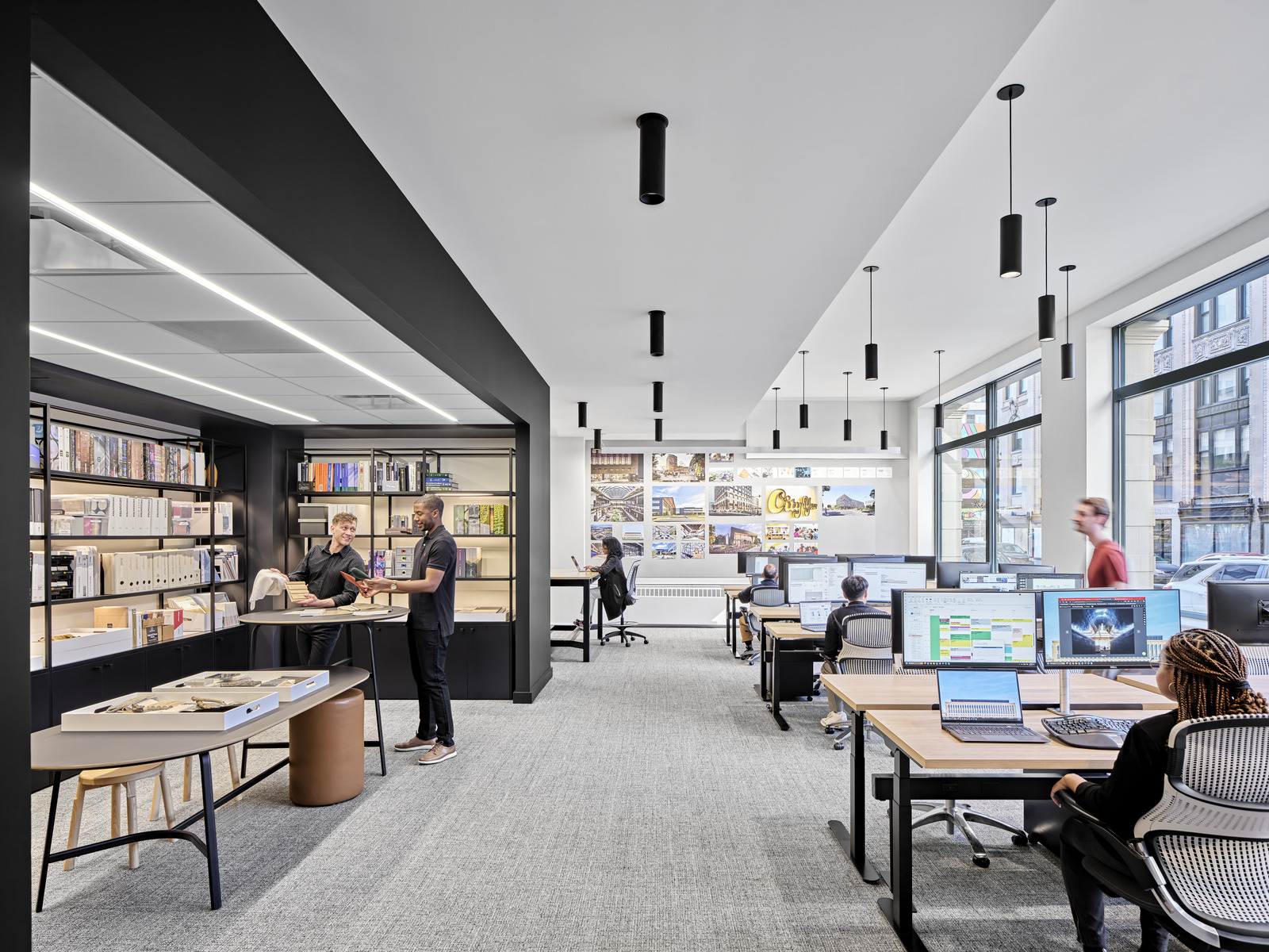 Modern open office with people working at desks and collaborating by shelves; large windows provide natural light, shelves display books and materials, and computer monitors show various projects.