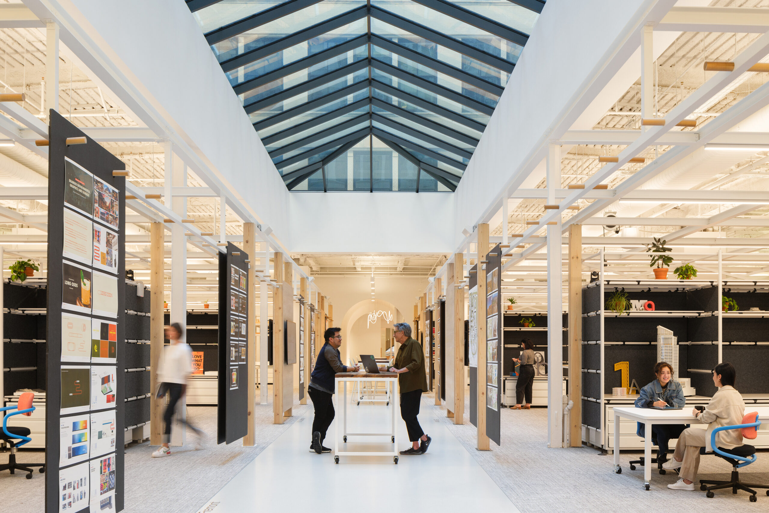 Modern open office space with a glass ceiling, two people standing at a mobile desk in the center, others working at tables and walking along the aisles.