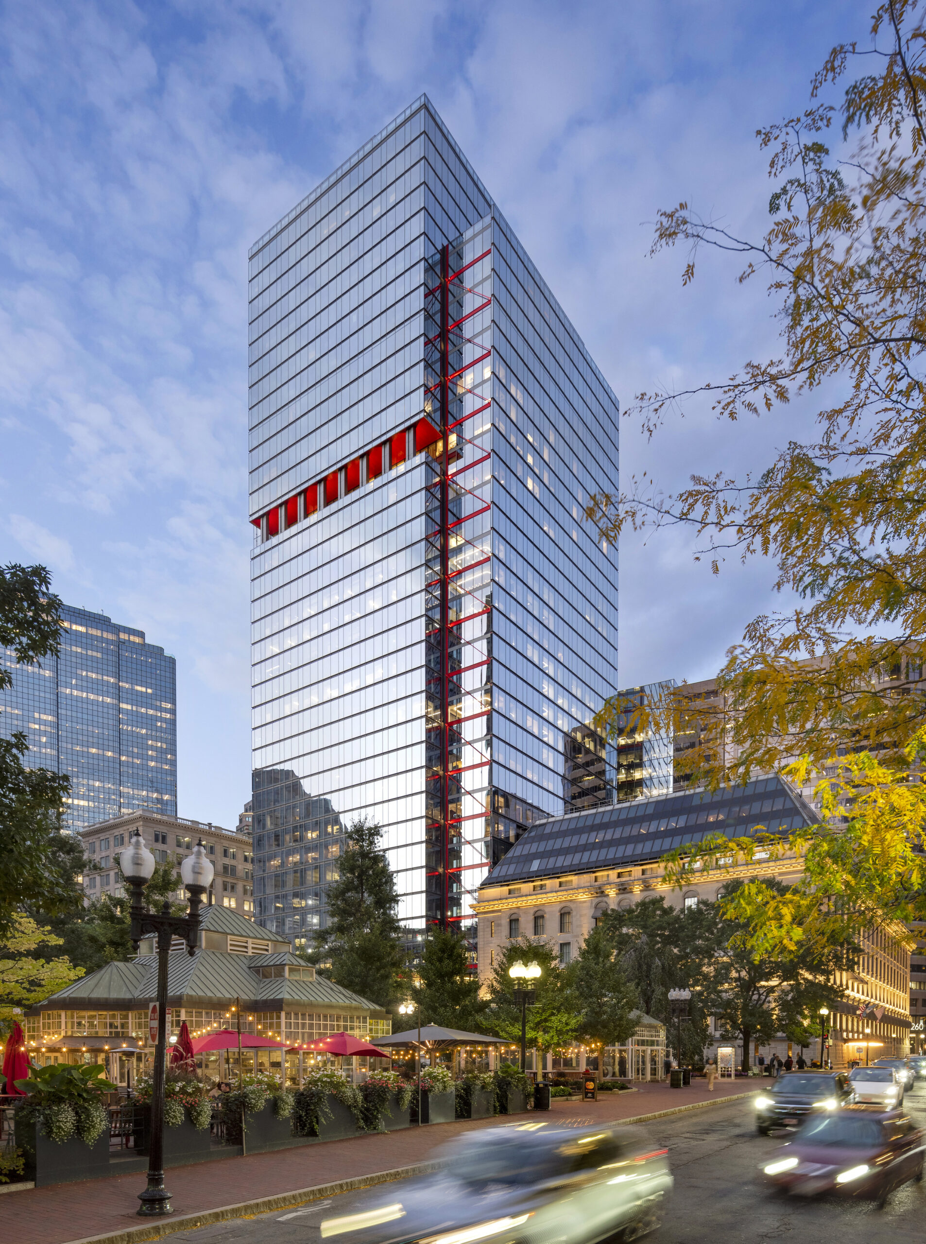 A modern glass office tower with red vertical accents, surrounded by trees and older buildings, stands above a busy city street at dusk.