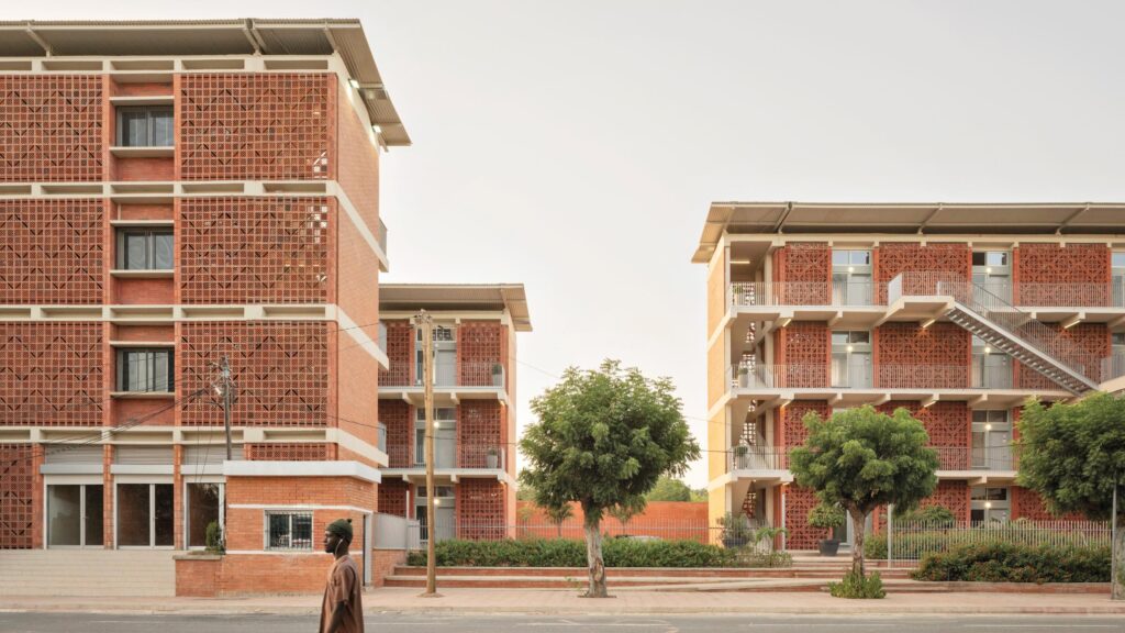 Two modern brick apartment buildings with patterned facades and exterior staircases stand beside a street lined with trees; a person walks in the foreground.