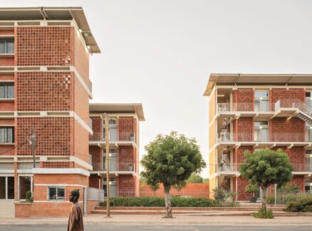 Two modern brick apartment buildings with patterned facades and exterior staircases stand beside a street lined with trees; a person walks in the foreground.