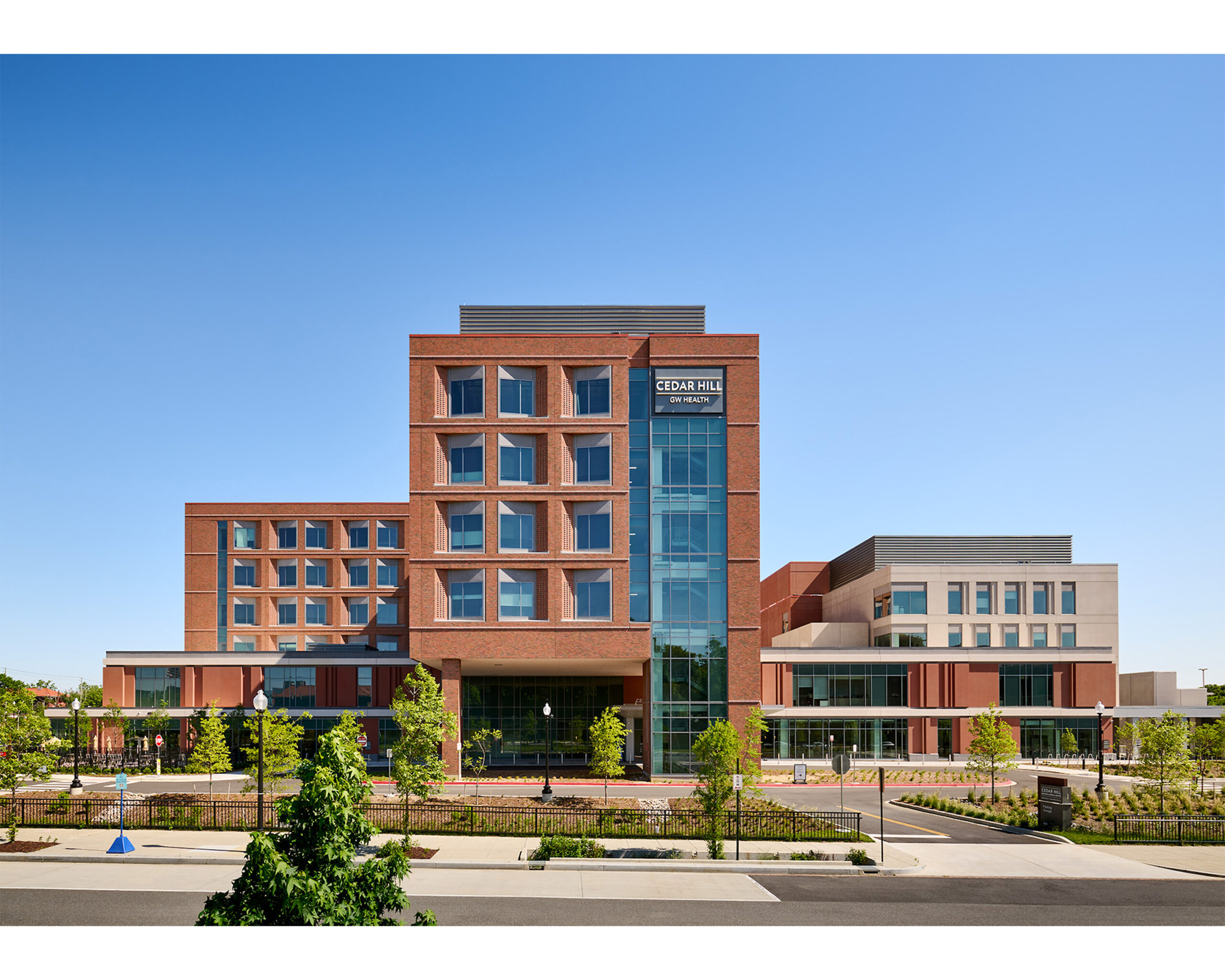 A modern, multi-story brick hospital building with large windows, glass entrance, and landscaped grounds under a clear blue sky.