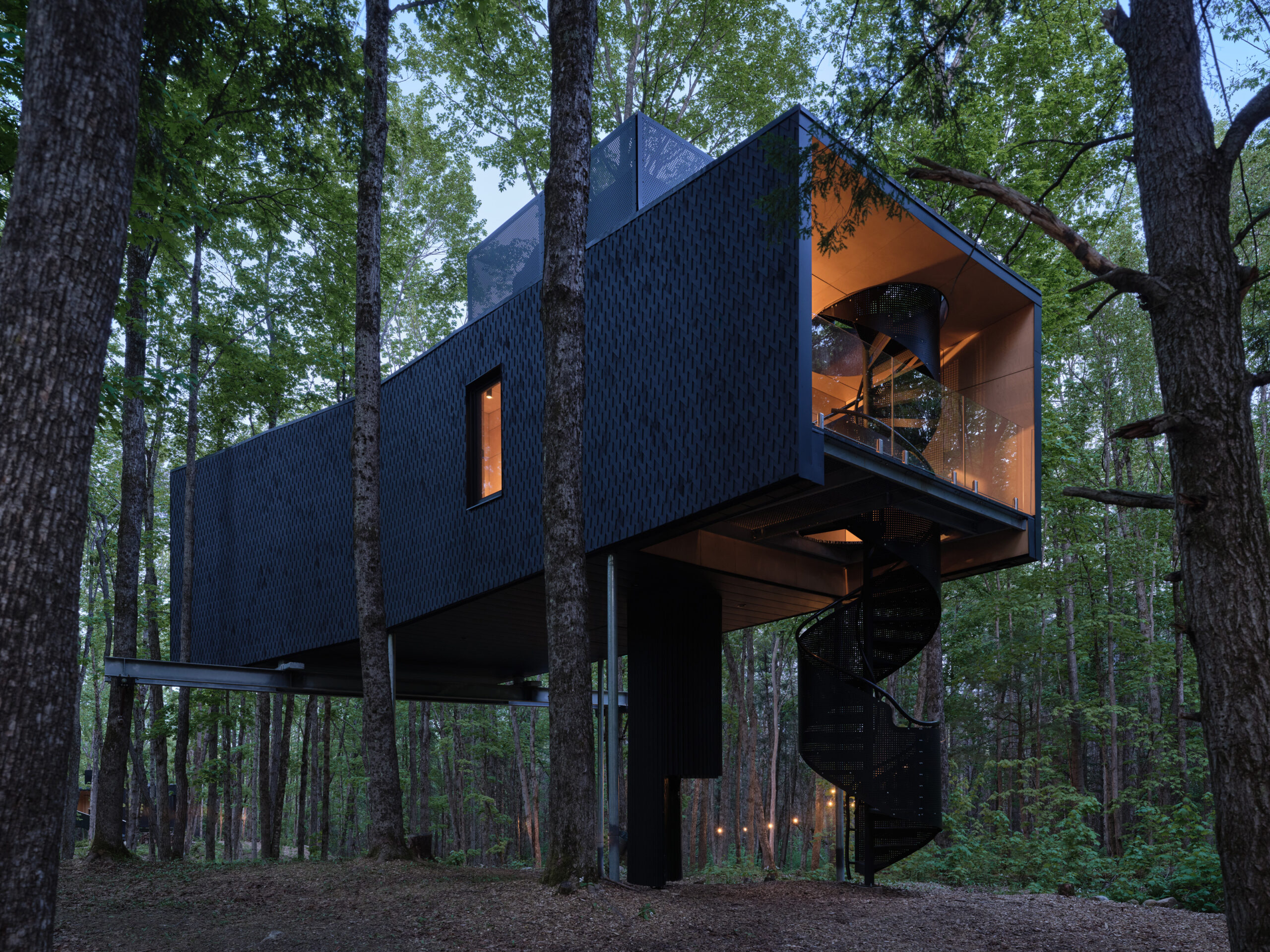 Modern black treehouse with geometric design, suspended among tall trees, featuring a spiral staircase and large glass window, set in a dense forest.