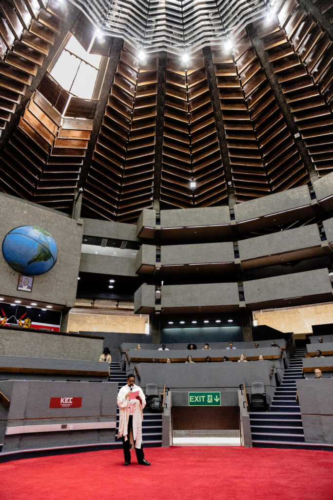 A person in a white coat stands on a red carpet inside a large, modern, circular building with tiered seating and a globe on the wall, evoking the innovative spirit of UIA 2026 Barcelona.