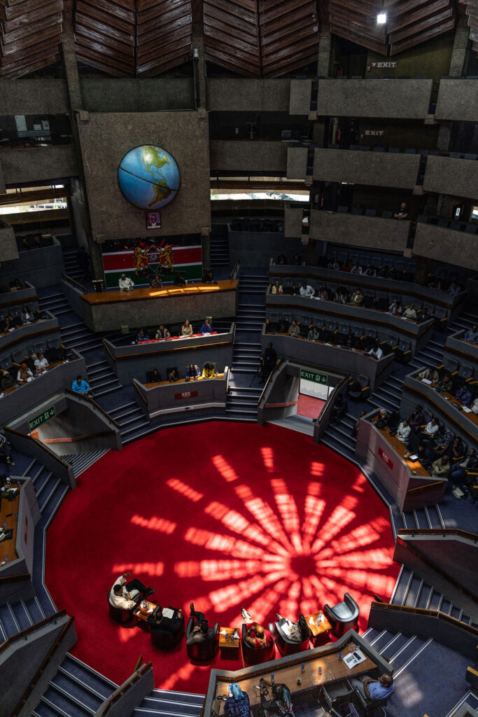 Large round legislative chamber with red carpet, people seated around circular desks, sunlight creating a patterned shadow on the floor, and a large globe—highlighting UIA 2026 Barcelona—displayed prominently on the wall above.