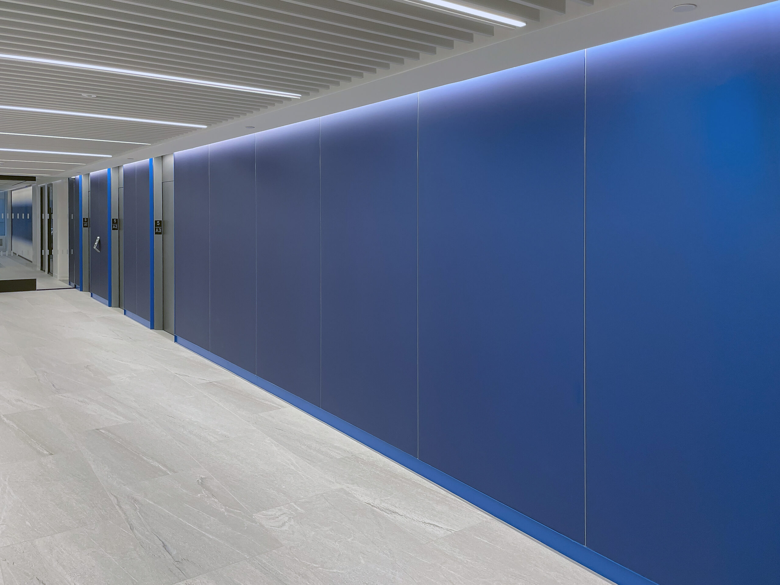 Modern hallway with blue wall panels, light-colored floor tiles, and a ceiling with linear recessed lighting. Elevator doors are visible on the left side of the hallway.